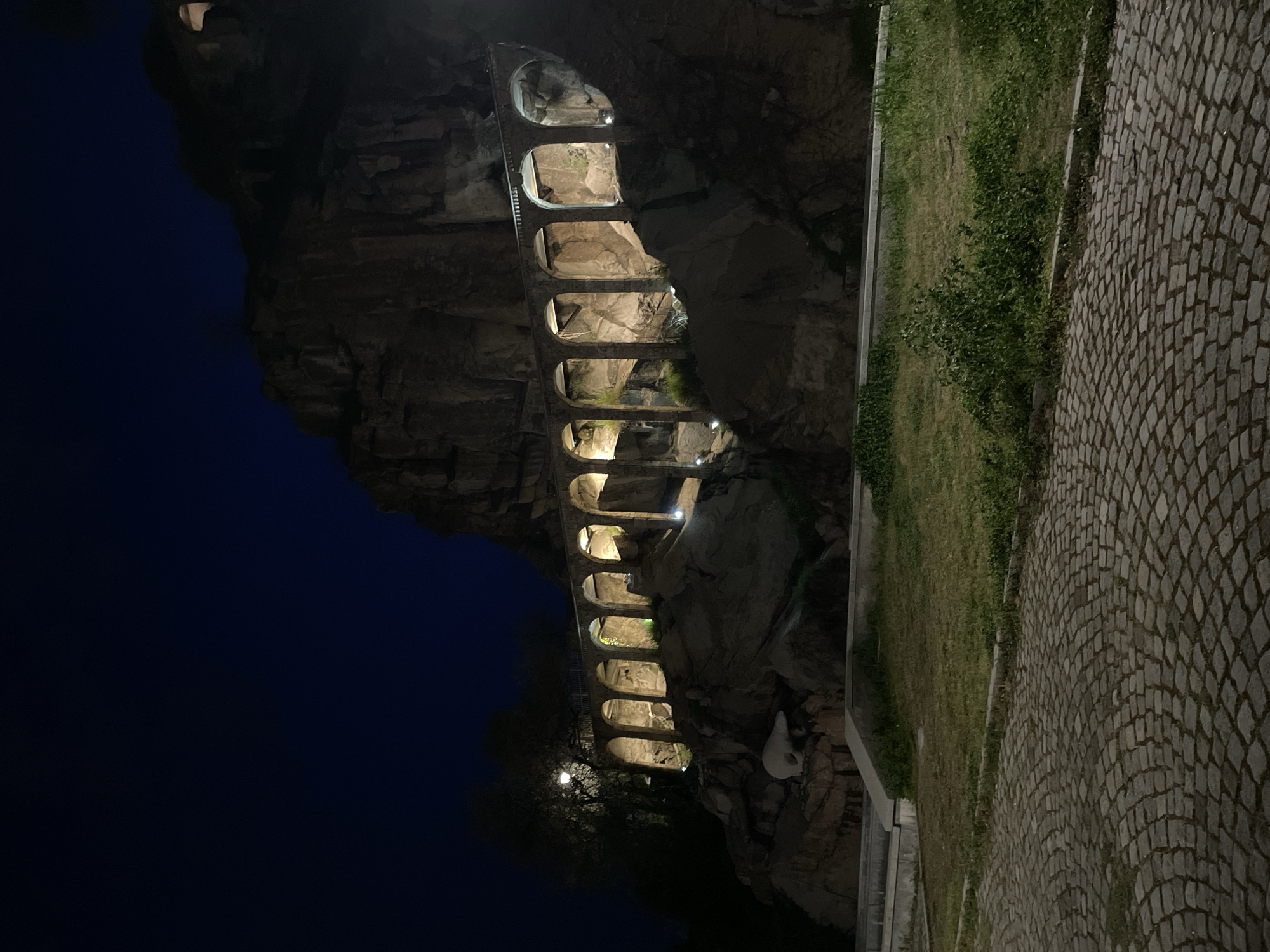 At night, a stone aqueduct in Plovdiv, Bulgaria, is lit. The aqueduct is built into a rocky cliff face and has a series of arches. Some of the arches are illuminated by lights. Next to the cliff is a grassy area and a cobblestone path. The sky is dark blue.