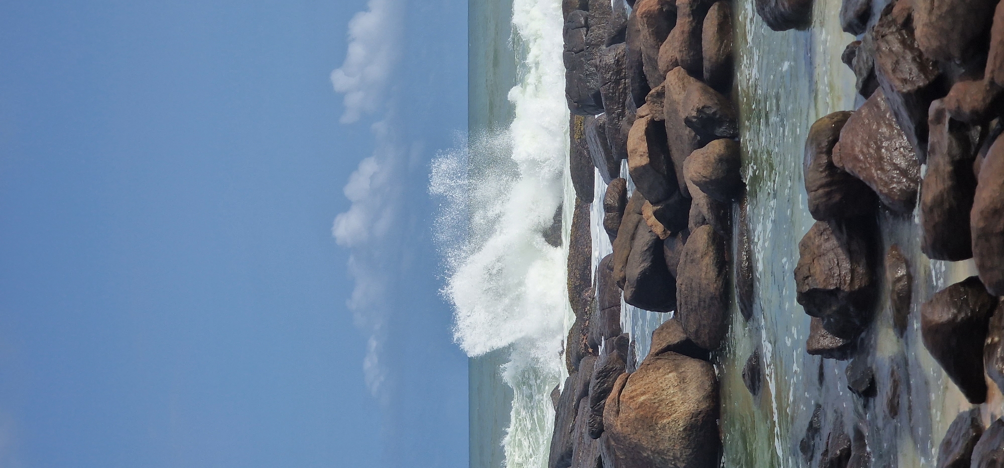 The image shows a rocky shoreline in Dondra, Sri Lanka. Large, dark rocks are clustered along the water's edge. A wave is crashing against the rocks, creating white spray. The sky is blue with scattered white clouds. The water is a mix of green and blue.