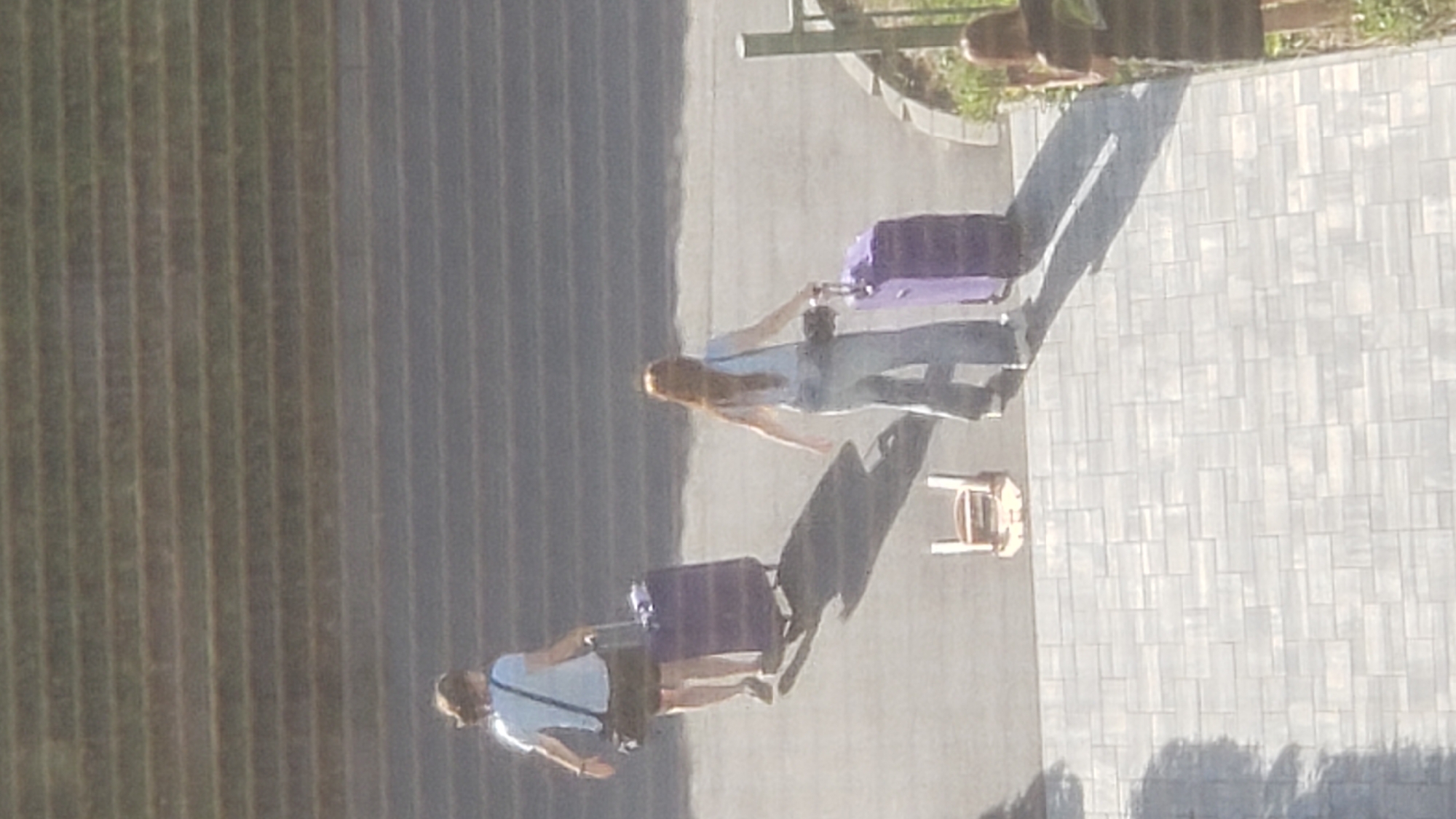 The image shows an aerial view of two individuals walking with luggage on an asphalt surface, with a background of horizontal steps. A third individual is partially visible on the right, facing away from the camera and holding a mobile phone. A small, light-colored chair is positioned on the asphalt between the two walking individuals. The ground in the foreground is paved with light gray rectangular tiles. The scene does not contain any identifiable landmarks or features that would allow for specific location determination within the city of Prešov, Slovakia.
