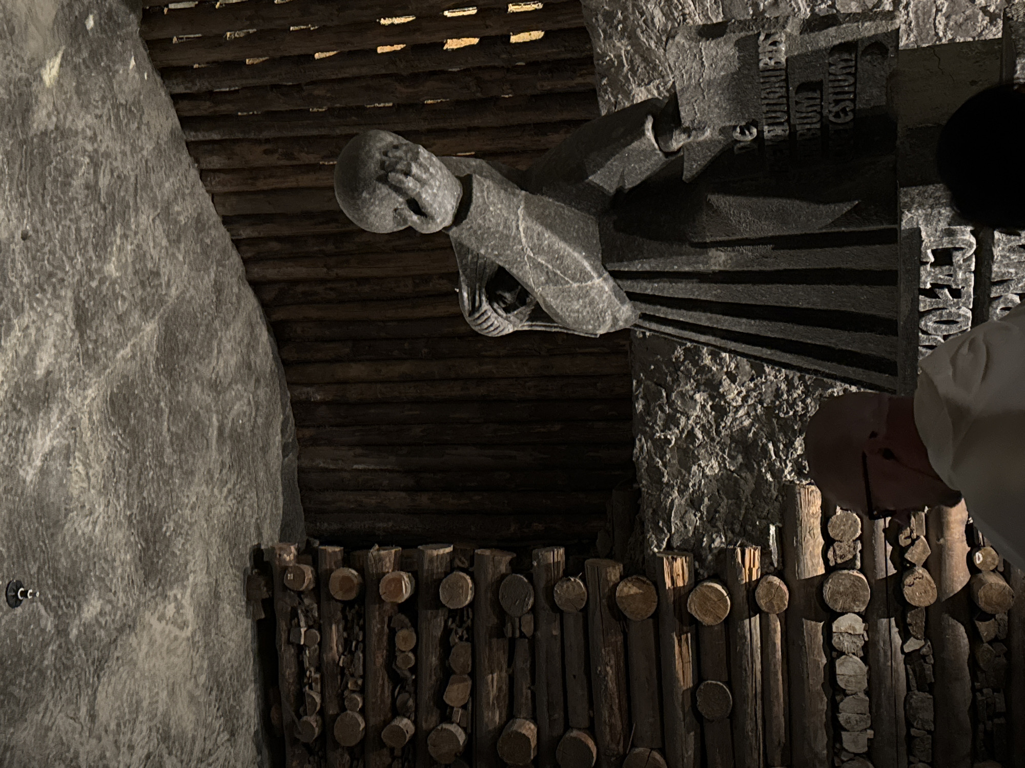 This is a rotated, low-angle, dimly-lit, indoor shot of a statue in Wieliczka, Poland.
The statue is a gray, carved figure of a monk with head in hand. To the right of the statue is a section of carved stone with lettering in an unknown language.
The back wall is made of dark logs and there is a railing along the bottom made of many smaller logs. A person in a white garment is standing in front of the log railing on the right. The person has dark hair. A rough, irregular-textured stone wall is on the left.