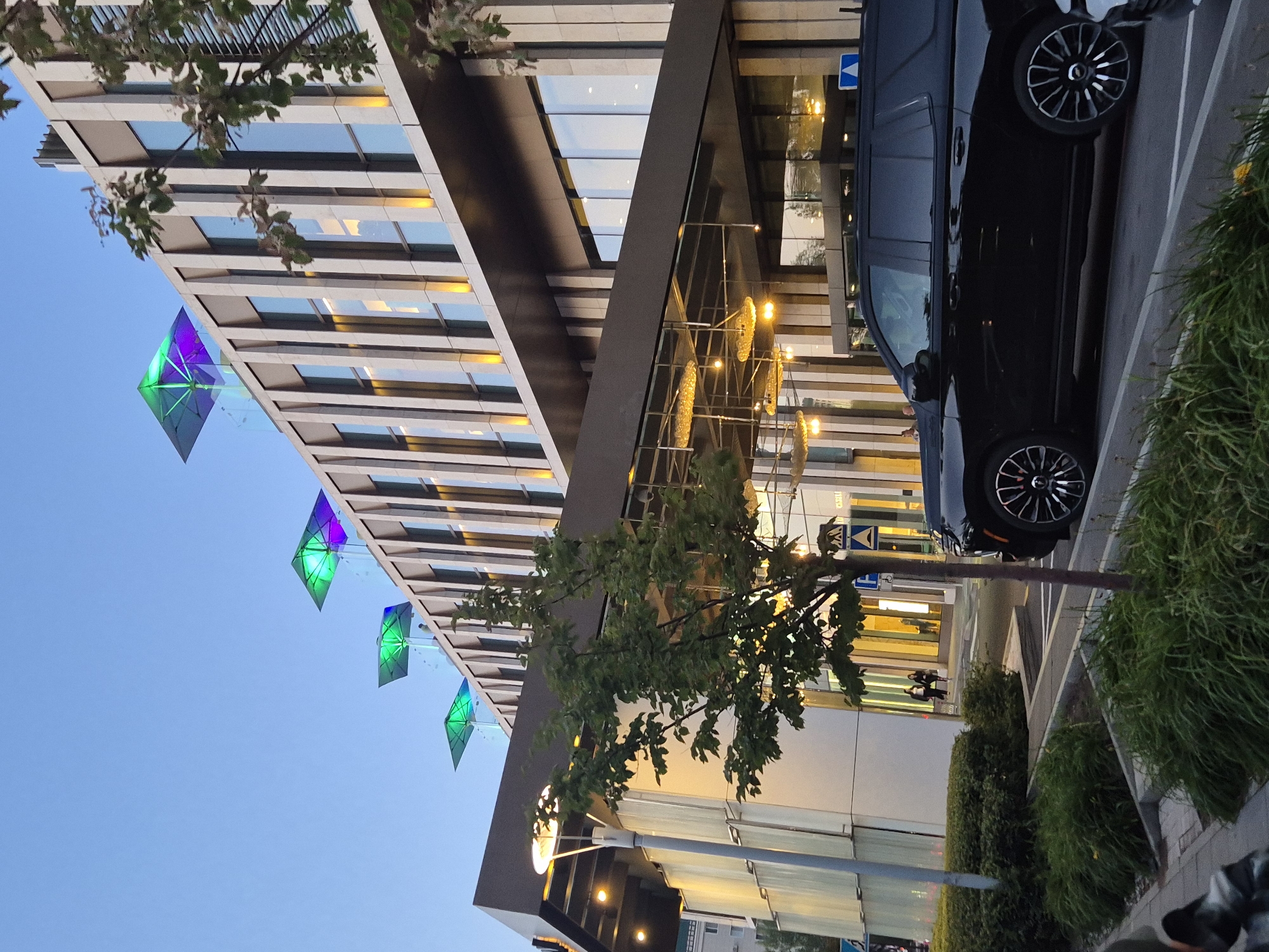 A black SUV is parked on the street in front of a modern, multi-story building in Astana, Kazakhstan. The building features a facade of vertical concrete elements and large glass windows, with some windows illuminated by warm yellow lights. On the rooftop, there are several square structures with illuminated green and purple lighting. A small tree with green leaves grows on the sidewalk in front of the building. The sky is clear and blue, indicating it is likely daytime. There are a few pedestrians visible near the entrance of the building. The street appears to be well-maintained, with clear lane markings and pedestrian crossings.