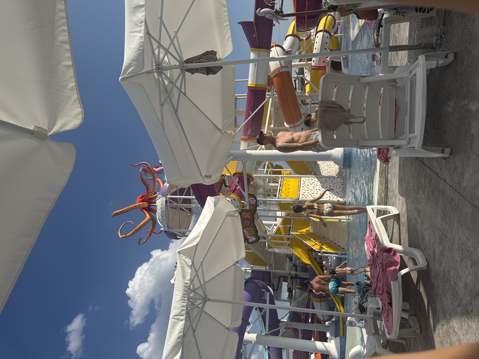 The image shows a water park on a sunny day. In the background, a large octopus-shaped slide structure is visible, with several colorful water slides descending from it. People are seen enjoying the water, with some in the pool and others on the slides. White umbrellas provide shade for beach chairs, some of which are occupied. A pink towel is draped over a lounge chair in the foreground. The ground is paved with concrete. The scene suggests a recreational area within a city, likely Ivanyane, Bulgaria, given the context.