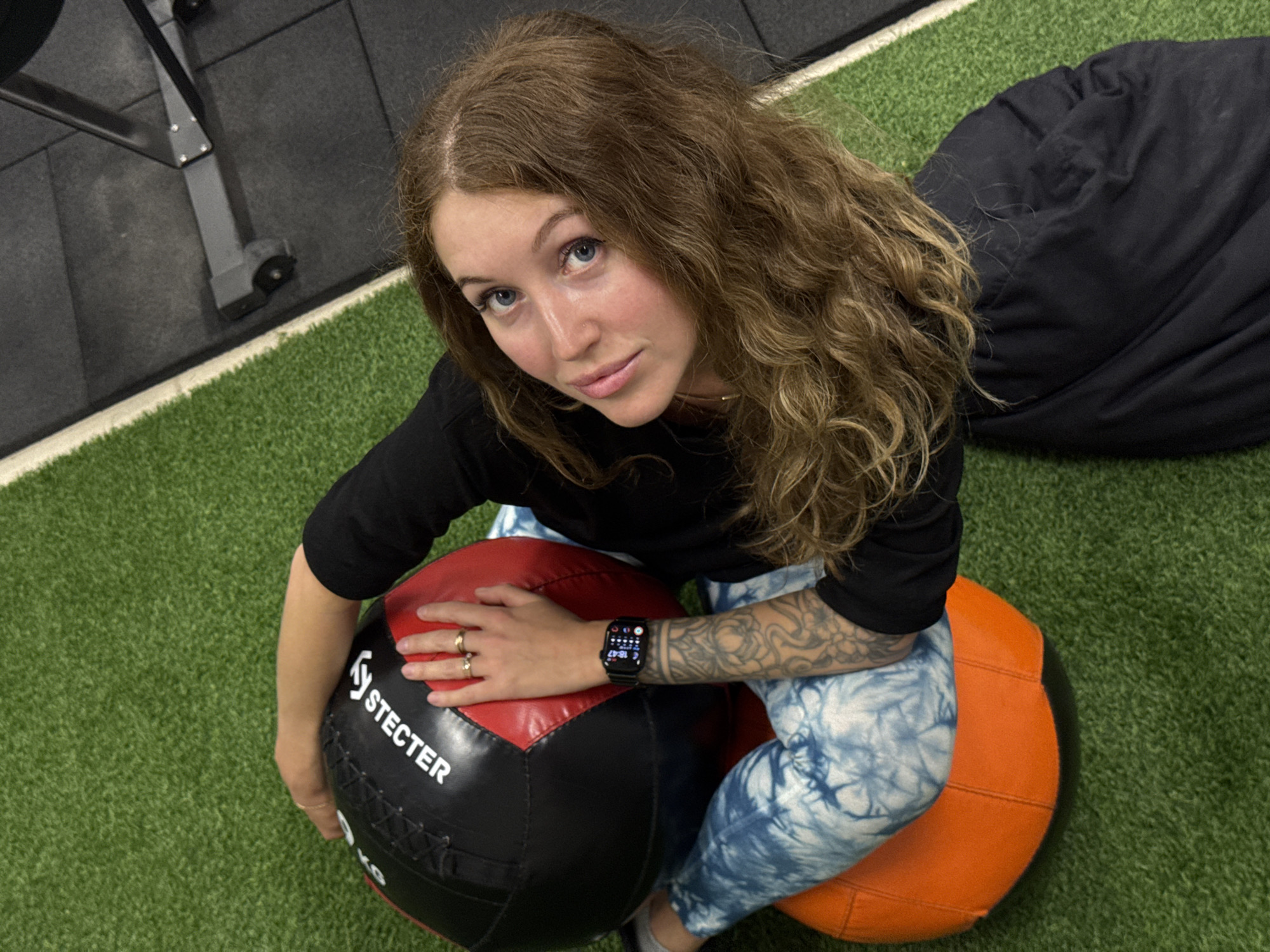 An overhead shot captures a young woman posing in an indoor gym or fitness facility in Chaikovsky, Russia. She has long, wavy brown hair and blue eyes, looking directly at the camera with a calm, inquisitive expression. She is seated on a large, orange and black weighted ball, with her hands resting on a red and black weighted ball in front of her. The red and black ball has "STECTER" printed in white text.
The woman is wearing a black long-sleeved top and blue tie-dye patterned leggings. On her left wrist, a smartwatch displays the time as 18:47, indicating the evening. Several rings adorn her right hand, and a prominent tattoo sleeve is visible on her left forearm. The floor of the facility features green artificial turf bordered by black rubber gym mats. Parts of black gym equipment and a dark bean bag or cushion are visible in the background. The overall scene suggests a posed photograph taken in a modern fitness environment.