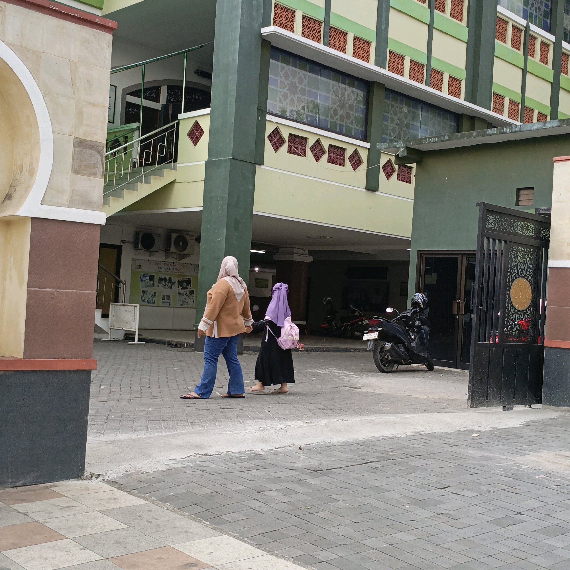 Two individuals, likely a mother and child, are walking on a paved walkway in front of a building. The person in front is wearing a tan jacket and blue jeans, while the person behind is wearing a purple hijab and a black dress. They are both wearing flip-flops. The building behind them appears to be a school or mosque, with green and light yellow exterior walls and ornate windows. There are air conditioning units mounted on the wall of the building. A motorcycle is parked near a black metal gate. The walkway is made of interlocking pavers. The scene is set in Salatiga, Indonesia.