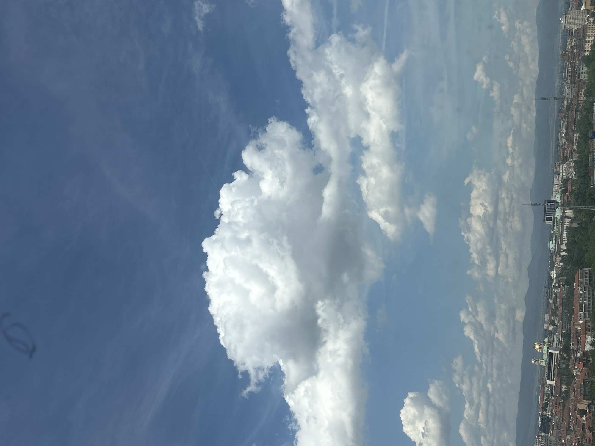 The image displays a panoramic view of a city beneath a bright, partly cloudy sky, with mountains in the distance. The sky, occupying the upper half of the frame, is a clear blue, dotted with various clouds. A large, dense, white cumulus cloud dominates the left-central portion of the sky, with other smaller, scattered white and gray cumulus clouds extending across the middle and right. Wispy cirrus clouds are faintly visible in the deeper blue sections of the sky. A faint, dark, irregularly shaped smudge, likely an optical artifact, is present in the upper left area of the sky.
The lower half of the image features a sprawling cityscape, indicative of Sofia, Bulgaria, as suggested by the location context. Buildings with diverse architectural styles are visible, many featuring reddish-brown tiled roofs. Green tree canopies are interspersed throughout the urban landscape. A notable structure with prominent golden domes can be seen within the city, consistent with the St. Alexander Nevsky Cathedral, a landmark in Sofia. Beyond the city, a range of hazy mountains, likely Vitosha Mountain, stretches across the horizon in the background.
The scene depicts a clear and sunny daytime with good visibility, under scattered cloud cover. No specific activity or event is taking place, and there is no visible text.