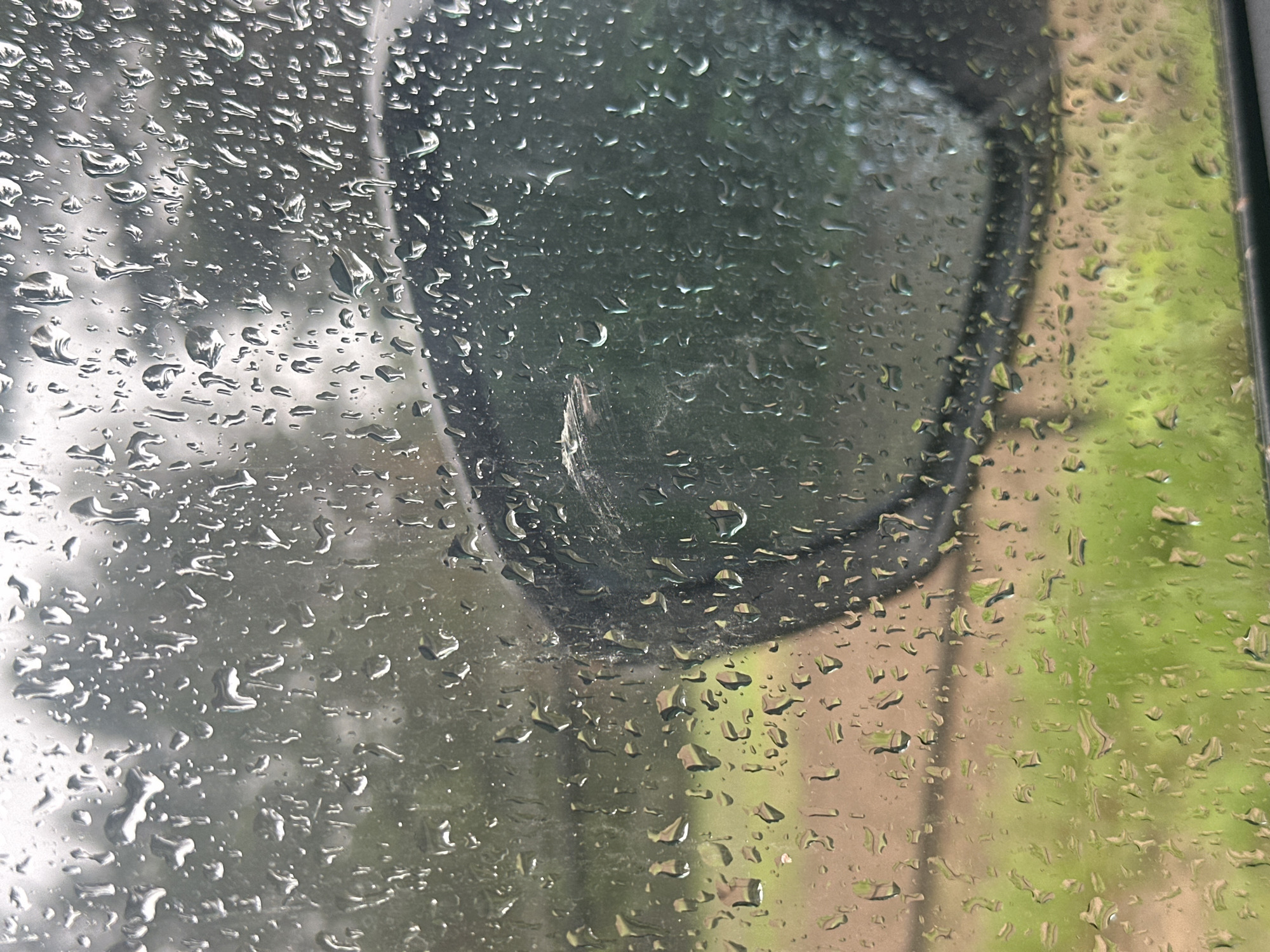 The image shows a rain-covered window, partially obscuring a side mirror of a vehicle. The view through the window reveals a blurred, likely outdoor scene with green foliage and possibly a building. The location appears to be in a city, due to the presence of the vehicle and surrounding structures, but the specific city is unknown. The focus of the image is the raindrops on the window, which distort the view behind it.