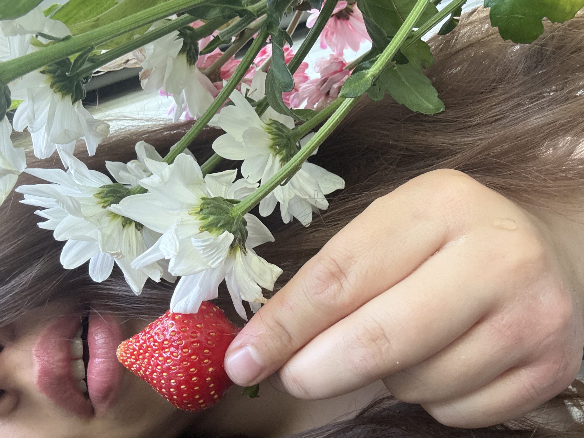 A close-up shot shows a person holding a strawberry and a bouquet of white and pink daisies. The person's mouth is visible in the upper left corner, with lips slightly parted. The strawberry is red and plump, with visible seeds, and is held between the index finger and thumb. The daisies are in various stages of bloom, with green stems and leaves. The background is blurred, with the person's brown hair dominating the right side of the frame. The lighting is soft and natural. The image does not contain any information that would allow for the determination of its location in Krasnoyarsk, Russia.