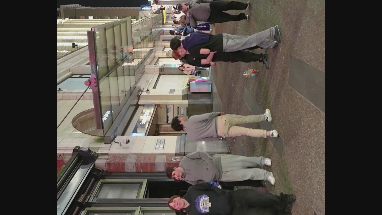 A group of young adults are gathered on a sidewalk in front of a building that appears to be a transit station, based on signage that reads "Granville Station." The architecture suggests an urban setting, likely Vancouver, Canada. In the foreground, four individuals stand on the sidewalk. To the far left, a young man with dark hair wears a black hoodie with a graphic print and dark pants. Next to him, a young man in a grey hoodie and light grey sweatpants stands with his hands in his pockets. To the right, a young man in a light grey long-sleeved shirt and light-colored cargo pants is facing away from the camera. Further to the right, two young men are engaged in a physical interaction, with one with red hair embracing the other, who is wearing a dark t-shirt and dark pants, and the latter has his arm around the former's shoulders. Another individual, a man with a dark baseball cap and a grey jacket, stands to the right, with his hand on the shoulder of another person. Several other people are visible in the background, some standing and others appearing to walk. Objects on the sidewalk include a colorful cup with a straw on the ground near the embracing individuals and what appear to be planters or decorative bins with colorful abstract designs. The building facade features large windows and an awning made of glass and metal. The lighting suggests it is evening or night.