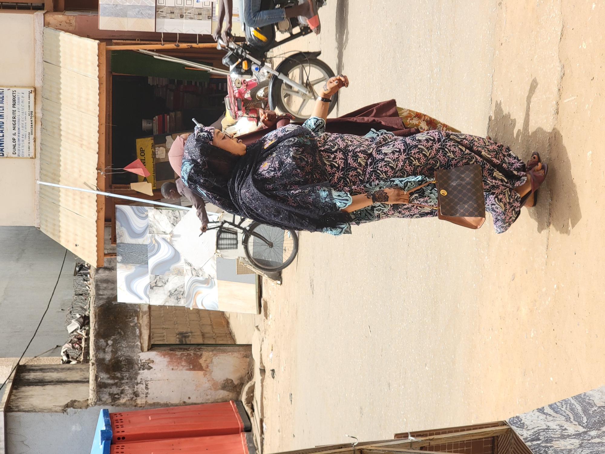 The image captures a street scene in what appears to be Maiburiji, Nigeria. A woman wearing a patterned dress and a black headscarf is walking on the sidewalk. She carries a brown purse. Next to her, a man is riding a bicycle, partially obscured by the woman. A motorbike is parked on the street. In the background, there is a store named "DUNKLAN INTER AGENCY," selling Dunlop and Nigerite products. The store has a metal roof and displays various tile samples. A sign indicates the store also sells tiles. A weathered building with brick and concrete construction is visible behind the store. The street itself is paved and shows signs of wear.