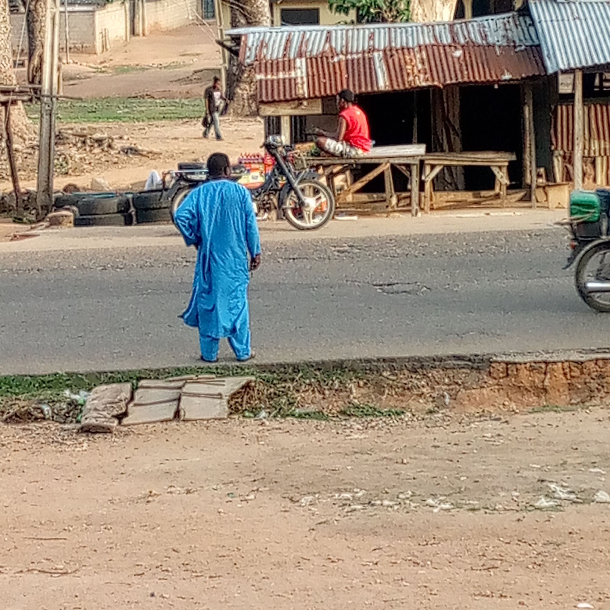This image captures a daytime scene along a worn asphalt road in what appears to be a rural or semi-urban setting. In the immediate foreground, facing away from the camera, a person dressed in a striking blue, long-sleeved tunic and trousers stands at the edge of the road, looking towards the right side of the frame.
The asphalt road runs across the middle of the image, showing cracks and potholes. On the opposite side of the road, beneath a makeshift shelter with a rusty, corrugated iron roof, another person is seated on a wooden table or stall. This individual wears a red t-shirt, camouflage shorts, and a black cap. A motorcycle is parked in front of this roadside structure.
Further in the background, a third person is seen walking on a dirt path, moving away from the viewer. To the left of the road, near the person in blue, a stack of old tires is visible. The ground bordering the road consists primarily of dry, light brown dirt interspersed with patches of green vegetation and some refuse. On the far left, what appears to be utility poles or tree trunks rise vertically. The overall scene depicts casual daily life and activity in an outdoor, somewhat informal environment. No text is visible in the image.