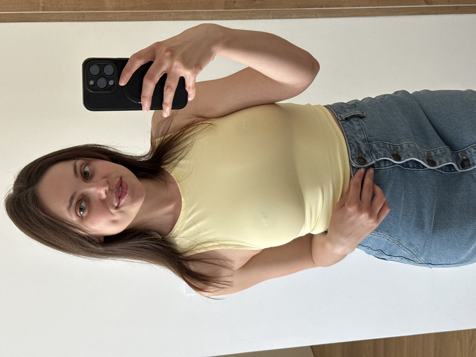 A woman is taking a selfie in front of a mirror. She is wearing a yellow tank top and a denim skirt with button detailing. She is holding a black iPhone in her right hand. The background is a white wall with a wooden trim along the top. It is impossible to determine the location in Belgrade, Serbia, from the image alone.