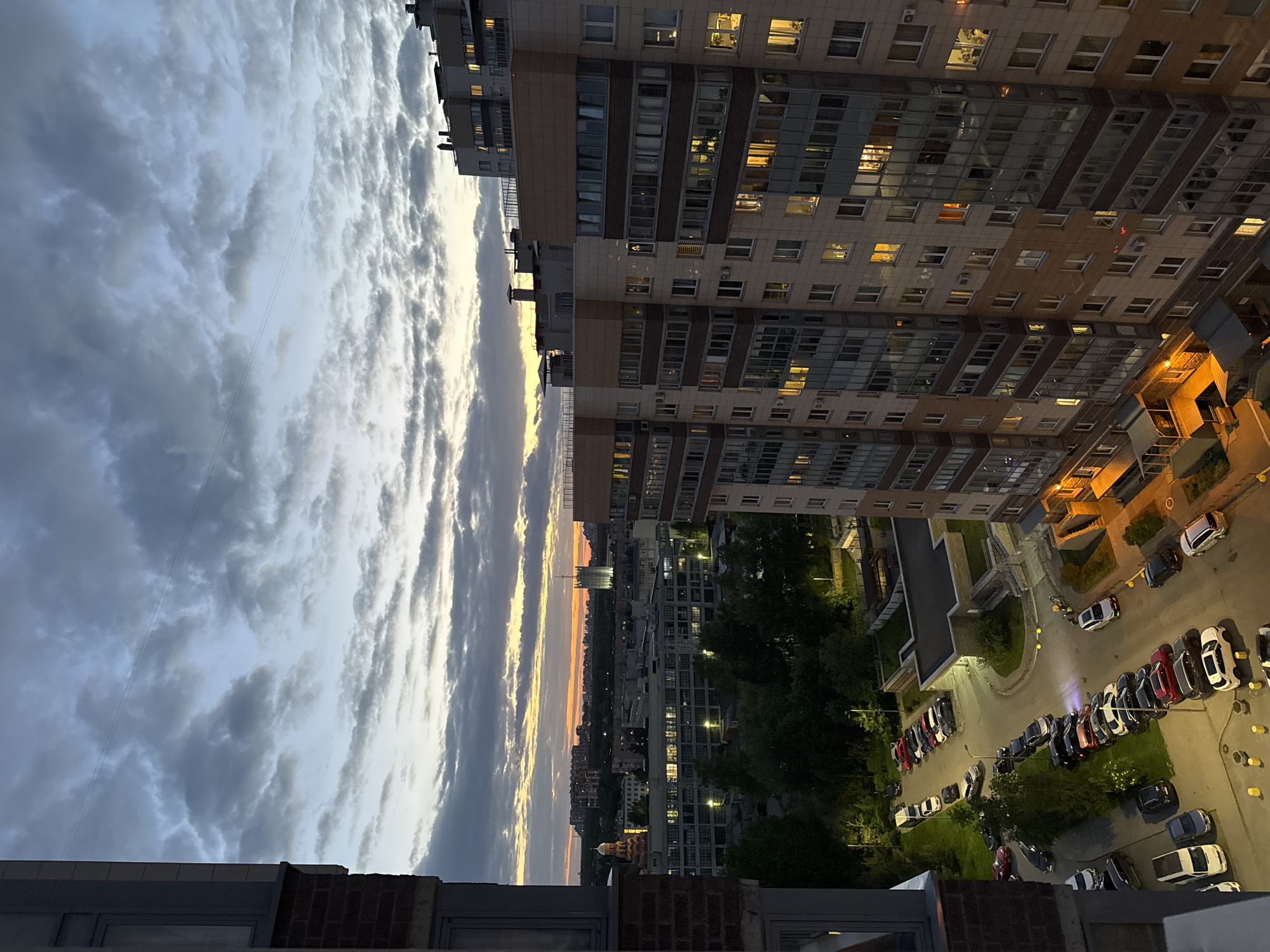 The image shows a view from a high vantage point, possibly a rooftop or upper floor of a building, overlooking an urban scene at dusk or twilight. The sky dominates the left side of the image, filled with clouds exhibiting varying shades of gray and white, suggesting a dynamic weather pattern. A strip of orange and yellow light indicates the position of the setting sun on the horizon.
On the right side, a tall, multi-story building is visible, with numerous illuminated windows suggesting residential occupancy. Below this building, a parking area is visible, filled with parked cars of various colors. Trees and green spaces are interspersed between the buildings and parking area. The general environment appears to be a residential area within a city. The specific location within the city of Unknown is not identifiable from the image. No people are visible in the image.