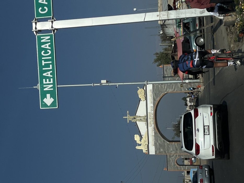 The image depicts a street scene in Nuevo Palmar, Mexico, under a clear blue sky. A large, ornate archway serves as a prominent landmark in the background, featuring a cross with a central figure, likely Christ, flanked by two relief sculptures, one resembling a church and the other an animal. Text above the arch reads "PARROQUIA SAN FELIPE NERI" and below it, "NUEVO PALMAR."
In the foreground, a male cyclist, viewed from behind, wears a multi-colored cycling jersey (blue, red, black, white), black cycling shorts, a helmet, and white socks. He is riding a red bicycle on the right side of the asphalt road, moving away from the viewer towards the archway. Immediately ahead of the cyclist is a white Kia Sportage SUV with a Mexican license plate "TXN-359-B," also proceeding in the same direction. A silver sedan is visible behind and to the left of the Kia.
To the right, a tall, light-colored utility pole supports a green directional sign with a left-pointing arrow, displaying the destination "NEALTICAN." Partially visible to the right of this sign is another green sign fragment. Beyond the cyclist and to the right, a green pickup truck is seen near various informal market stalls. Several other individuals, some on bicycles and others on foot, are visible near the archway and market area, indicating pedestrian and market activity. Small plants or flowers are arranged on the ground at the far right edge of the frame.