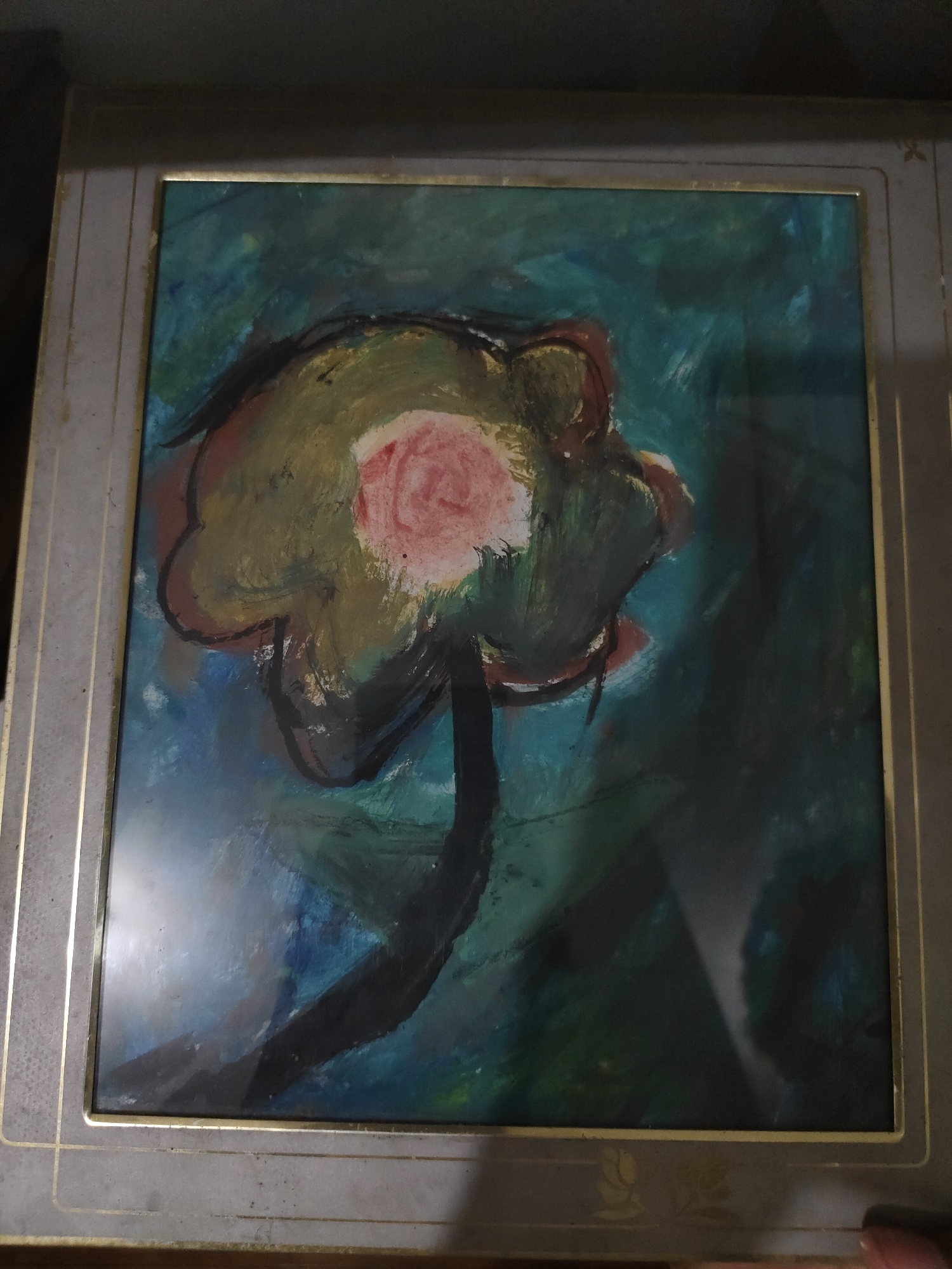 A framed painting depicting an abstract flower is shown from a slightly elevated perspective. The artwork features a large, stylized bloom centrally positioned, characterized by an irregular, organic outline in dark brown or black. The flower's petals are rendered in broad strokes of muted olive green, yellow, and reddish-brown, with a distinct, brighter pink-red circular center. A thick, curved stem in dark brown or black extends downwards from the base of the flower. The background of the painting is a mix of dark blues and greens, rendered with visible brushstrokes, creating an atmospheric effect.
The painting is encased in a wide, rectangular frame of a dark grey or charcoal color. The frame has thin, gold-colored lines accentuating its inner and outer edges. Faint, decorative gold motifs, possibly stylized floral or scroll patterns, are visible along the bottom section of the frame. The painting is covered by a reflective surface, likely glass or acrylic, which shows reflections from an external light source, particularly prominent in the lower left and bottom right quadrants of the image. The immediate environment surrounding the frame is dark and indistinct, providing no specific contextual details beyond the framed artwork itself. The scene is located in the city of Unknown, Unknown.