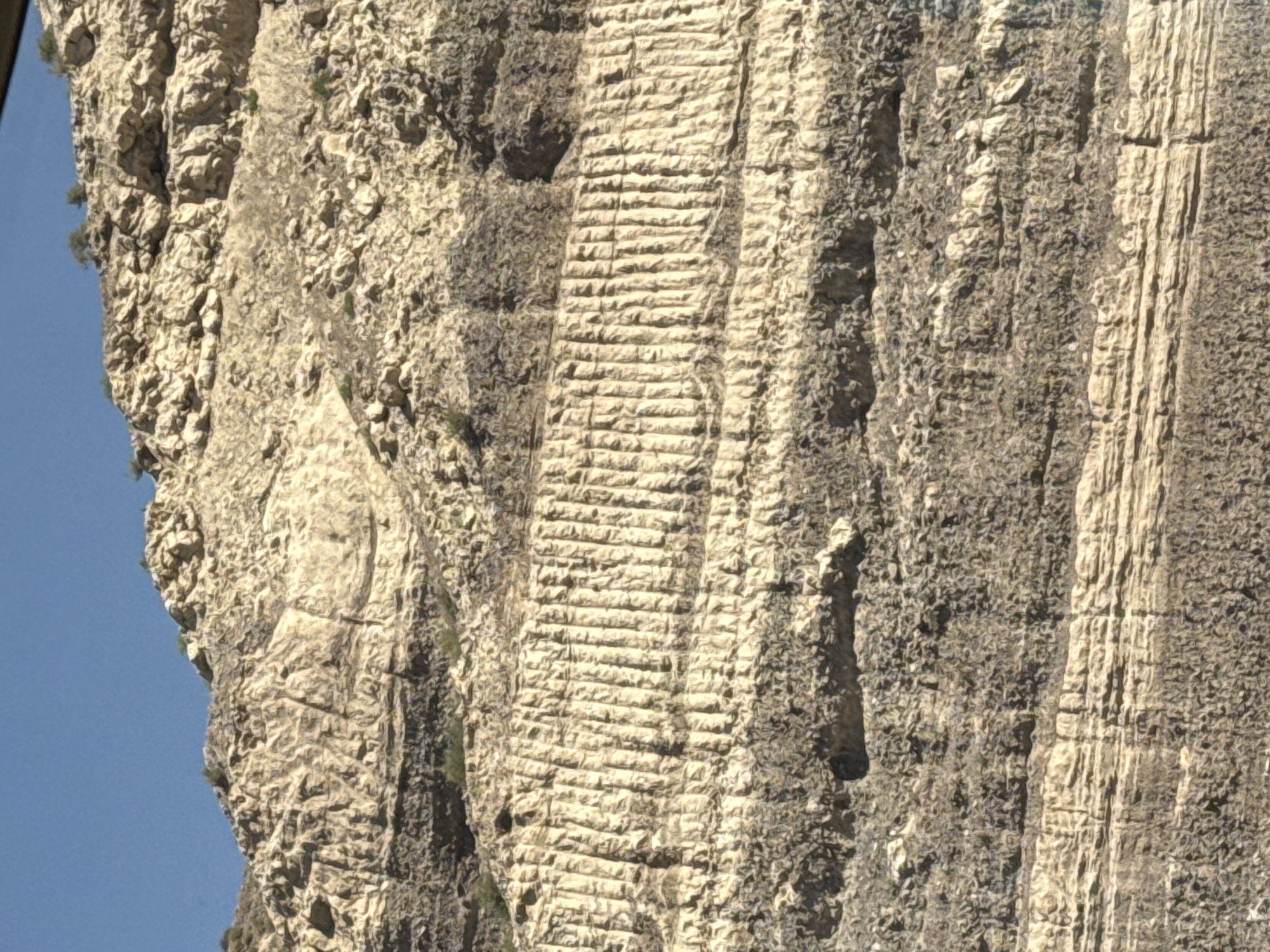The image shows a close-up view of a rocky hillside under a clear blue sky. The hillside is composed of distinct horizontal layers of sediment, with variations in color and texture. Some layers are light-colored and appear to be composed of fine-grained material, while others are darker and more granular. There are also visible vertical lines etched into one of the lighter colored layers, resembling grooves or striations. Sparse, dry vegetation, appearing as small green tufts, is scattered across the hillside, particularly on the upper slopes. The overall impression is one of geological stratification and erosion. The scene is devoid of people or any man-made structures.