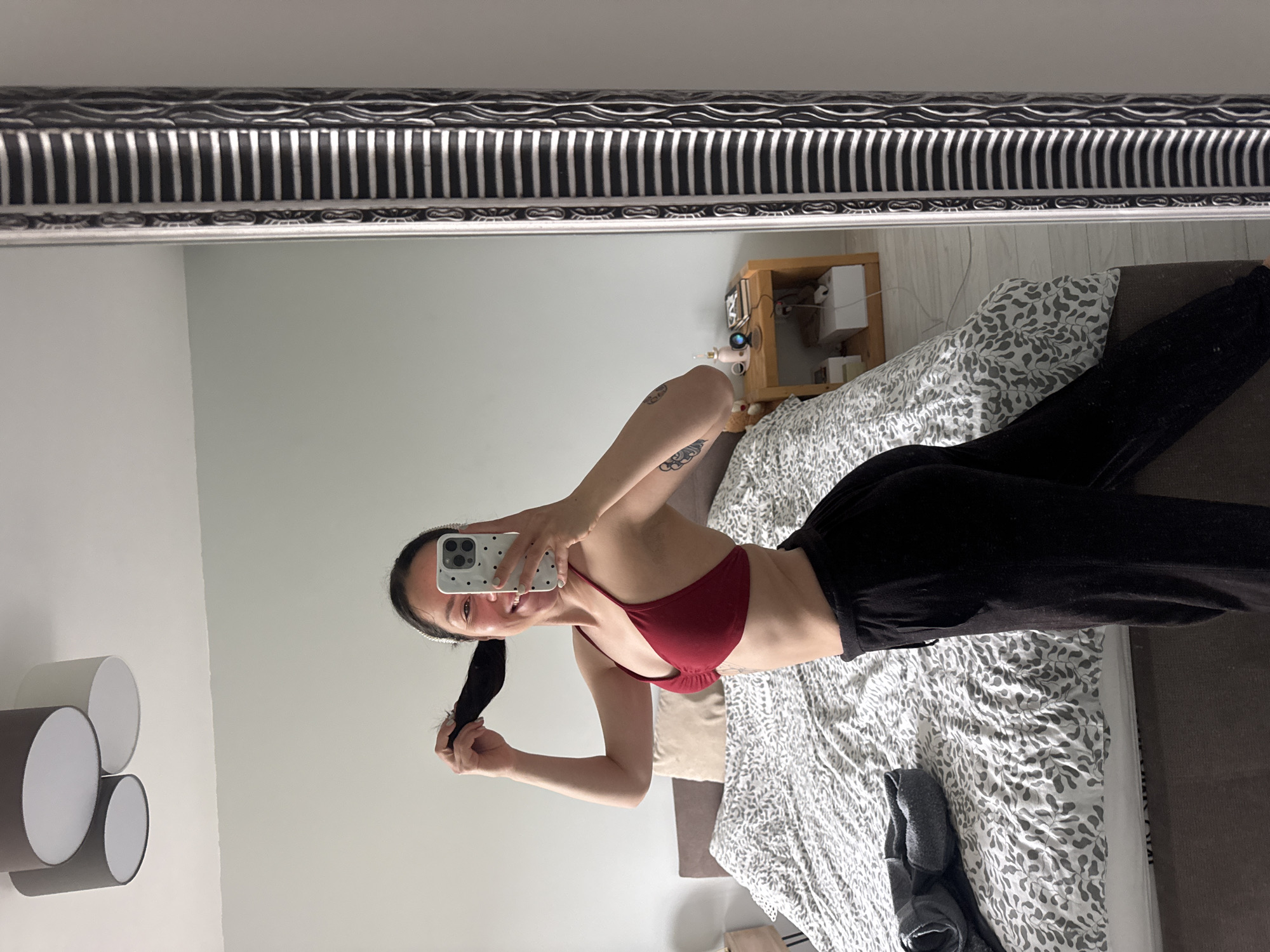 A young adult female is captured in a mirror selfie, standing in what appears to be a bedroom. She is smiling broadly and holding her dark hair, which is styled in a ponytail. She wears a red bralette or sports top and black pants. Tattoos are visible on her left arm. The person is holding a white smartphone with a black dotted or patterned case, through which the selfie is taken.
The scene is reflected in a large mirror with an ornate, silver-colored frame featuring intricate patterns along its edges. The background reveals a tidy bedroom setting. To the right, a bed is made with a white duvet cover patterned with grey, leaf-like motifs. A wooden bedside table with various small objects, including what looks like a lamp and a tissue box, is visible next to the bed. On the opposite wall from the bed, a modern wall-mounted light fixture with two cylindrical shades (one white, one grey) is visible. The walls are painted a light, neutral color. The overall lighting is bright and even, suggesting daytime. The person appears happy and relaxed. The location context provided is Burgas, Bulgaria. There is no visible text in the image.