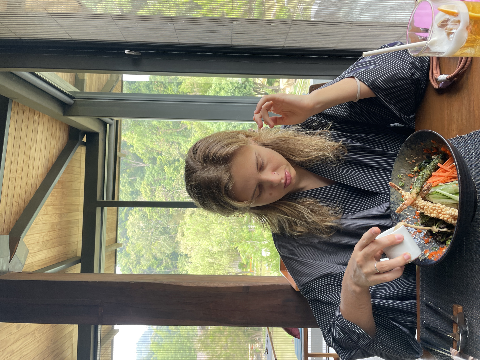 A woman is seated at a table next to a large window in a structure with wood and metal beam architecture. The view outside is a lush, green landscape. She is wearing a striped top and has light brown hair. In front of her is a bowl of food containing fried items, vegetables, and other ingredients. She is holding a small white cup over the bowl, possibly preparing to add sauce or dressing. To the upper right of the frame, there is a glass of liquid with ice and a straw. The photograph was taken in Ban Nong Hoi, Thailand.