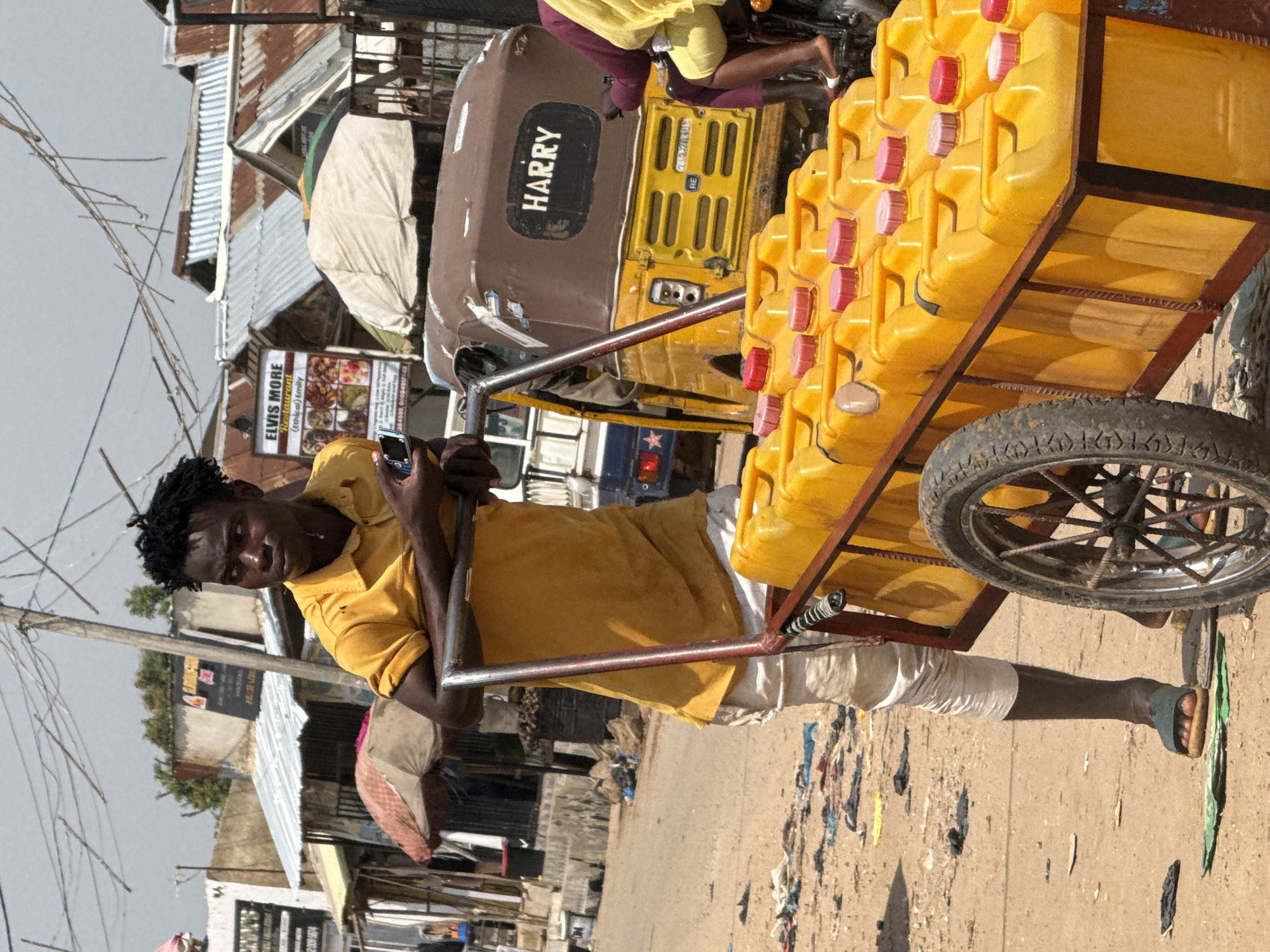 The image depicts a street scene in Abuja, Nigeria, under a bright, overcast sky. In the foreground, a young Black man with dark, curly hair is standing next to a cart loaded with numerous yellow plastic jerrycans, each capped with a red or pink lid. He is wearing a mustard-yellow polo shirt and light-colored pants, with dark sandals on his feet. His arms are crossed, and he is holding a mobile phone in his right hand. The cart itself is made of dark metal with two wheels, one of which is prominently visible.
Behind the man and the cart, a three-wheeled vehicle, likely an auto-rickshaw, is parked. It has a brown canopy and "HARRY" is visible in white lettering on the side. The background reveals a bustling market or street area with several buildings featuring corrugated metal roofs, some of which appear weathered. Umbrellas and awnings suggest stalls or shops. The ground is unpaved and appears dusty, with scattered debris including plastic bags and other refuse. Utility poles and wires are visible against the sky, indicating an urban environment. A person in a purple garment and yellow head covering is partially visible on the right, possibly riding on the back of a motorcycle. The overall impression is of a busy, informal commercial setting.