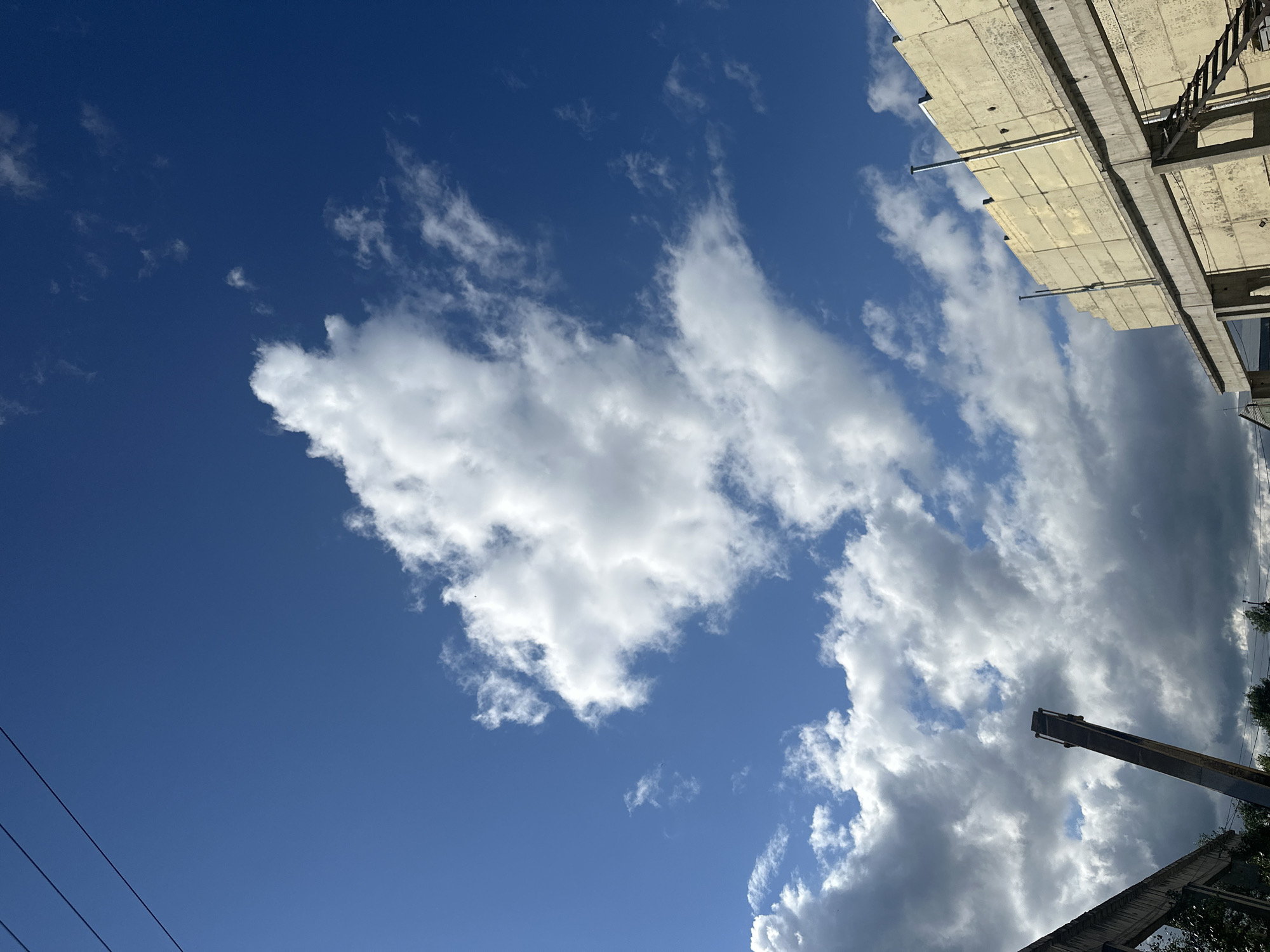 This media file captures a low-angle view of a clear blue sky filled with fluffy white clouds. In the upper right corner, the edge of a concrete structure, possibly under construction or renovation, is visible. It features horizontal slabs and vertical support beams, with some signs of wear and tear. A metal ladder is attached to one of the beams. In the lower right, a dark, angular object, likely part of construction equipment such as a crane boom, extends upwards against the cloudy sky. The lower left of the frame shows three dark, horizontal lines, which are likely power lines. The overall scene suggests an outdoor setting, possibly a construction site, on a bright day with scattered clouds. The orientation of the image is such that the sky occupies the majority of the frame, with the construction elements framing the top and right edges. There is no visible text in the image. The time of day appears to be daytime, likely late morning or afternoon, given the bright sunlight illuminating the clouds and the clear blue sky. The weather seems pleasant with a mix of sun and clouds.