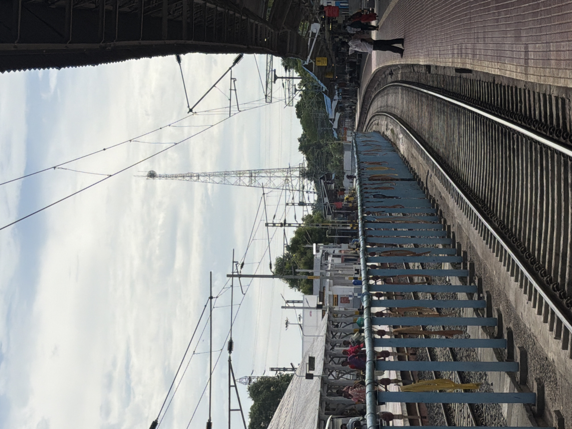 The image depicts a busy train station platform in Thanjavur, India. Several people are visible on the platform, some standing and others appearing to be walking. The scene is set outdoors under a cloudy sky, suggesting it is daytime.
Dominant objects in the scene include train tracks, overhead electrical wires, and a tall metal tower in the distance, likely a communication tower. A blue metal railing with wooden planks runs alongside the tracks, separating the platform from the tracks themselves. Various structures that appear to be part of the station's infrastructure, such as pillars and sheds, are also present. The platform surface is paved with reddish-brown bricks.
The composition of the image, with its emphasis on the curved train tracks and the overhead wires, creates a sense of depth and activity. The presence of people suggests a normal day at the station, with commuters or travelers possibly waiting for trains or disembarking. The lighting indicates it is likely midday or afternoon, with diffused sunlight due to the clouds. There is no visible text in the image.