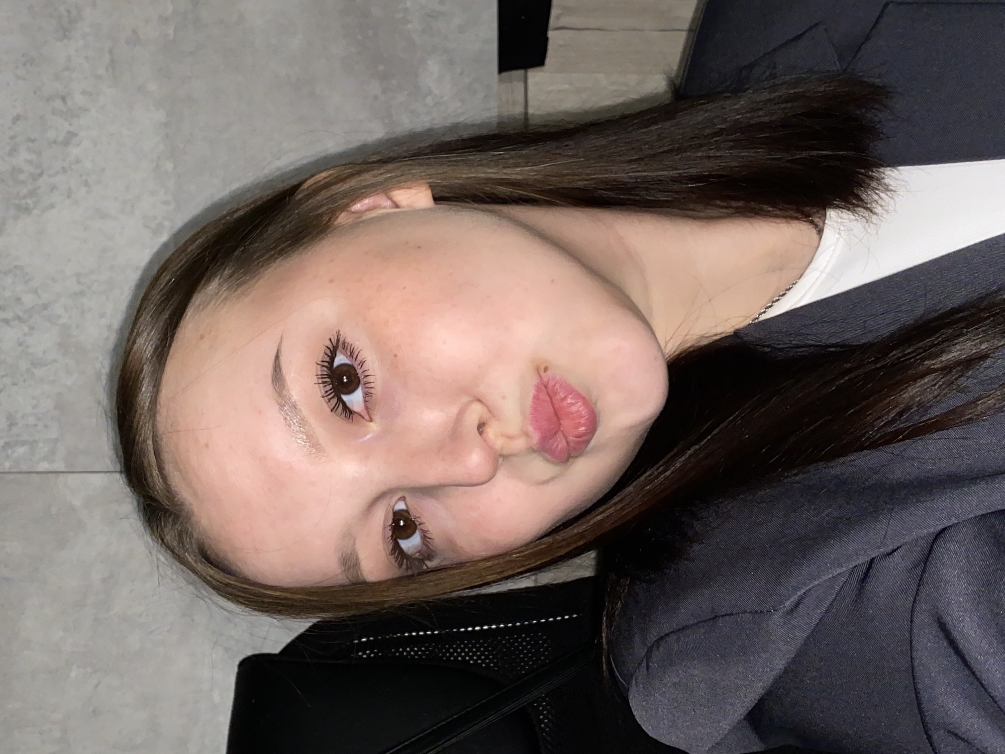 A young woman with brown hair and eyes is posing for a photo. She has fair skin with some freckles and is making a kissing face. She is wearing a dark-colored blazer over a white top. The background appears to be a neutral-colored wall and a black object, possibly part of a chair or luggage. The location is not identifiable as Chaikovsky, Russia based on the provided visual information.
