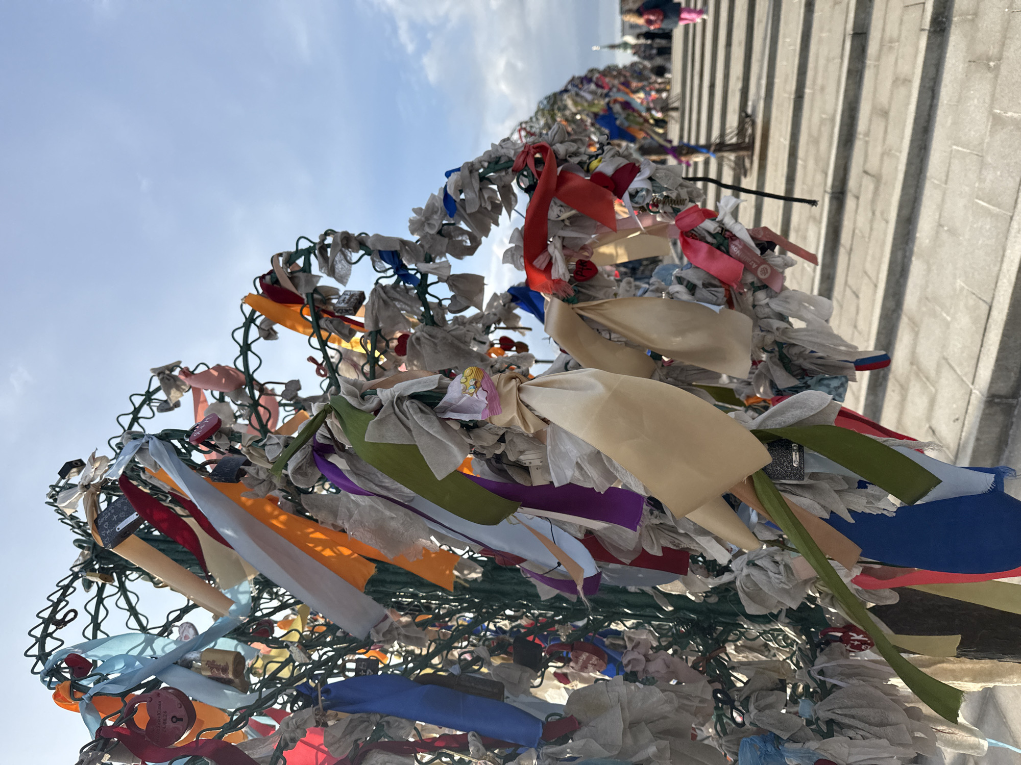 The scene depicts a section of a "love tree" on the Luzhkov Bridge (also known as Tretyakov Bridge or Kissing Bridge) in Moscow, Russia. The foreground is dominated by a green, wrought-iron sculptural 'tree' heavily laden with a dense accumulation of multi-colored fabric ribbons and numerous small padlocks. Ribbons are present in various hues including blue, orange, red, white, beige, purple, and green, tied in knots and bows. Padlocks are of diverse shapes, including heart-shaped, and some exhibit visible inscriptions such as '10 лет' (10 years) and '10.08.24'. The structure is one of a series visible along a paved pedestrian walkway that recedes into the background, where additional identical 'trees' are present. The overhead sky is clear blue with some scattered clouds. Several indistinct human figures are visible on the distant walkway, including one individual wearing a blue coat and pink pants on the right.