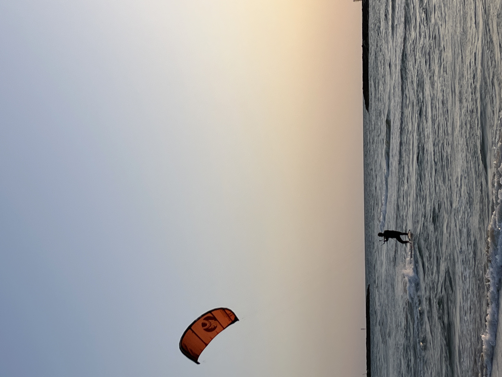 An orange kitesurfing kite is visible against a gradient sky. A person is kitesurfing in the ocean near a shore. The location appears to be an unknown city in an unknown region.
