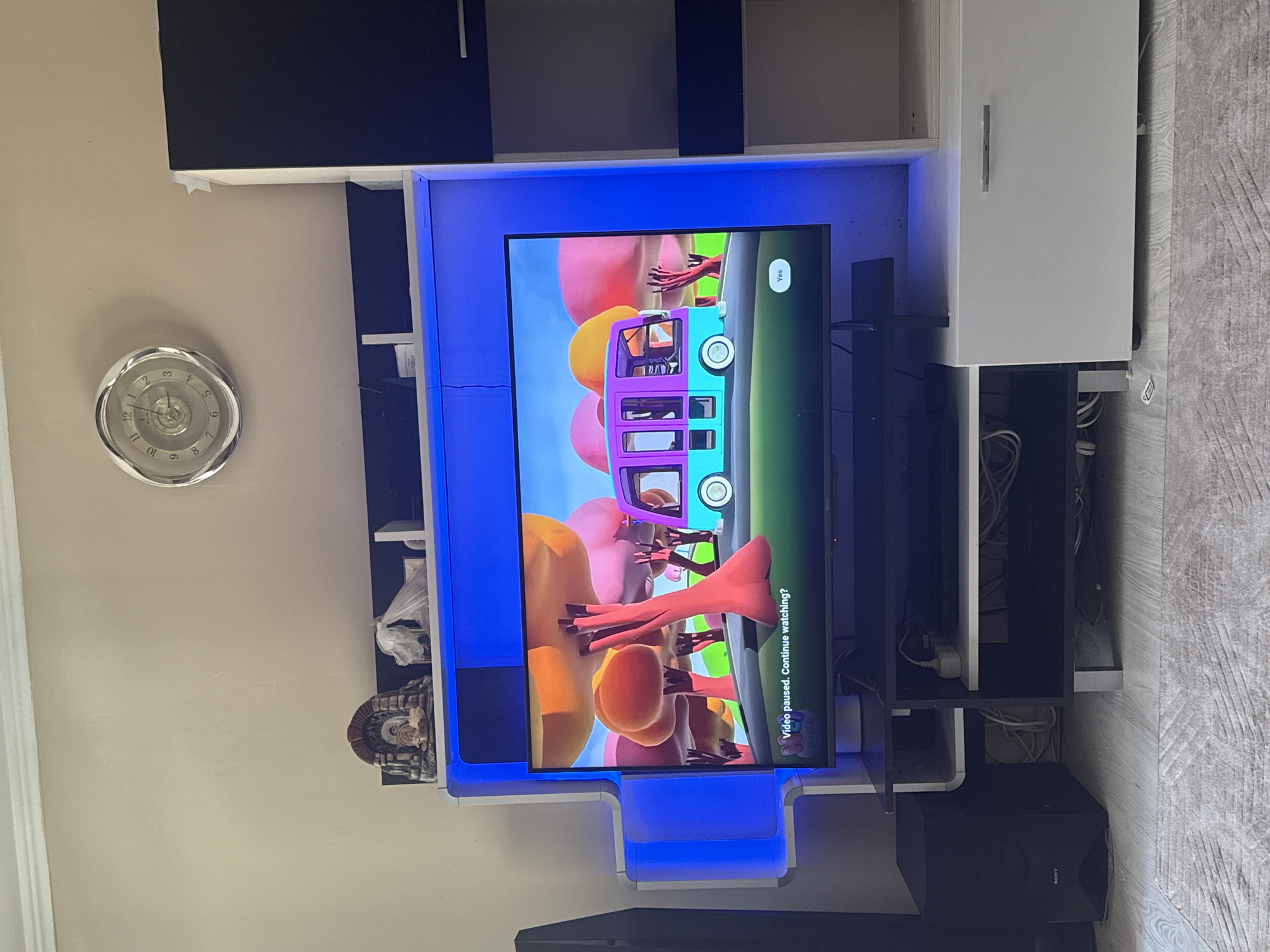 The image shows a modern entertainment unit with a large television screen displaying a paused children's cartoon. The cartoon features bright, colorful characters, including a bus and what appear to be abstract figures or objects, possibly balloons. The television is framed by blue LED lighting, contributing to a contemporary ambiance.
To the left of the television, a silver-toned clock with Roman numerals is mounted on a light-colored wall. The clock's design is intricate, with a decorative bezel. Below the clock, on the wall, is a small, decorative object that resembles a miniature shrine or statue with a rounded top.
The entertainment unit itself is a mix of dark and light-colored cabinets and shelves. Some shelves are empty, while others contain various items, including what appears to be a white plastic bag and some coiled wires, suggesting electronic equipment or cables are stored there. A black subwoofer with the "SONY" logo is positioned on the floor to the right of the entertainment unit.
The overall setting appears to be a living room or media room. The lighting is somewhat dim, with the blue light from the television unit being a prominent source of illumination. The scene is static, with the video paused, indicating a moment of rest or interruption. The visible text on the screen reads "Video paused. Continue watching?" and a "Yes" button, common on streaming platforms. The "SONY" logo on the subwoofer confirms the brand of some of the electronics. The clock suggests it is not nighttime, but the precise time is difficult to determine due to the angle and resolution of the clock face.