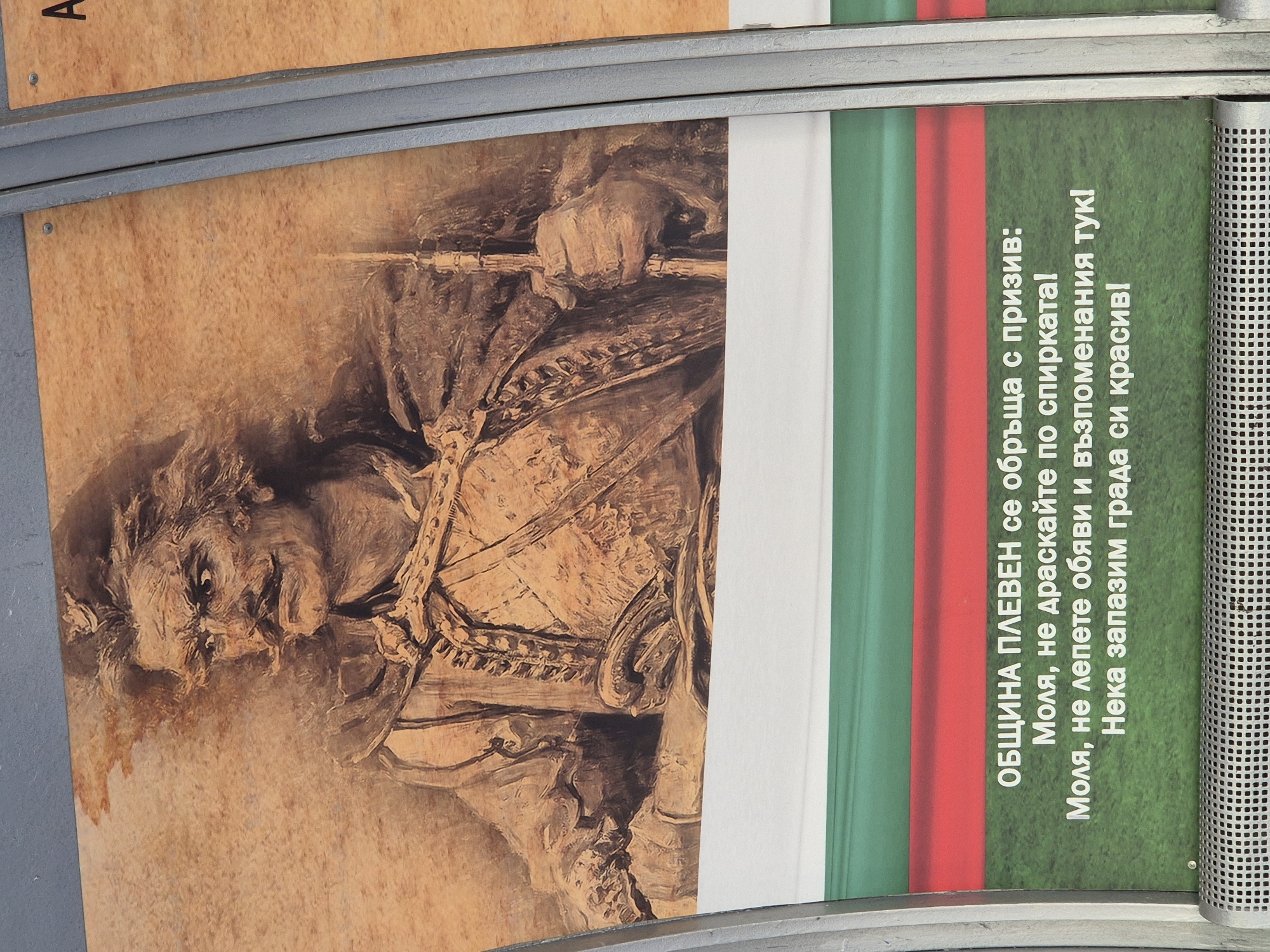 The image shows a close-up of a poster featuring a historical figure's portrait and text in Bulgarian. The portrait, rendered in a sepia tone, depicts a man with a prominent mustache, wearing traditional attire and holding a staff. The background of the poster is a textured, wood-like pattern. Below the portrait, there are horizontal stripes of white, red, and green, resembling the Bulgarian flag. The text below the stripes is printed in white against a green background. The text reads: "ОБЩИНА ПЛЕВЕН се обръща с призив: Моля, не драскайте по спирката! Моля, не лепете обяви и възпоменания тук! Нека запазим града си красив!" which translates to "Municipality of Pleven appeals: Please do not scratch the bus stop! Please do not post notices and memorials here! Let us keep our city beautiful!". The poster is framed by metal structures, suggesting it is displayed outdoors, possibly at a bus stop given the text. A portion of another poster with the letter "A" is visible to the right.