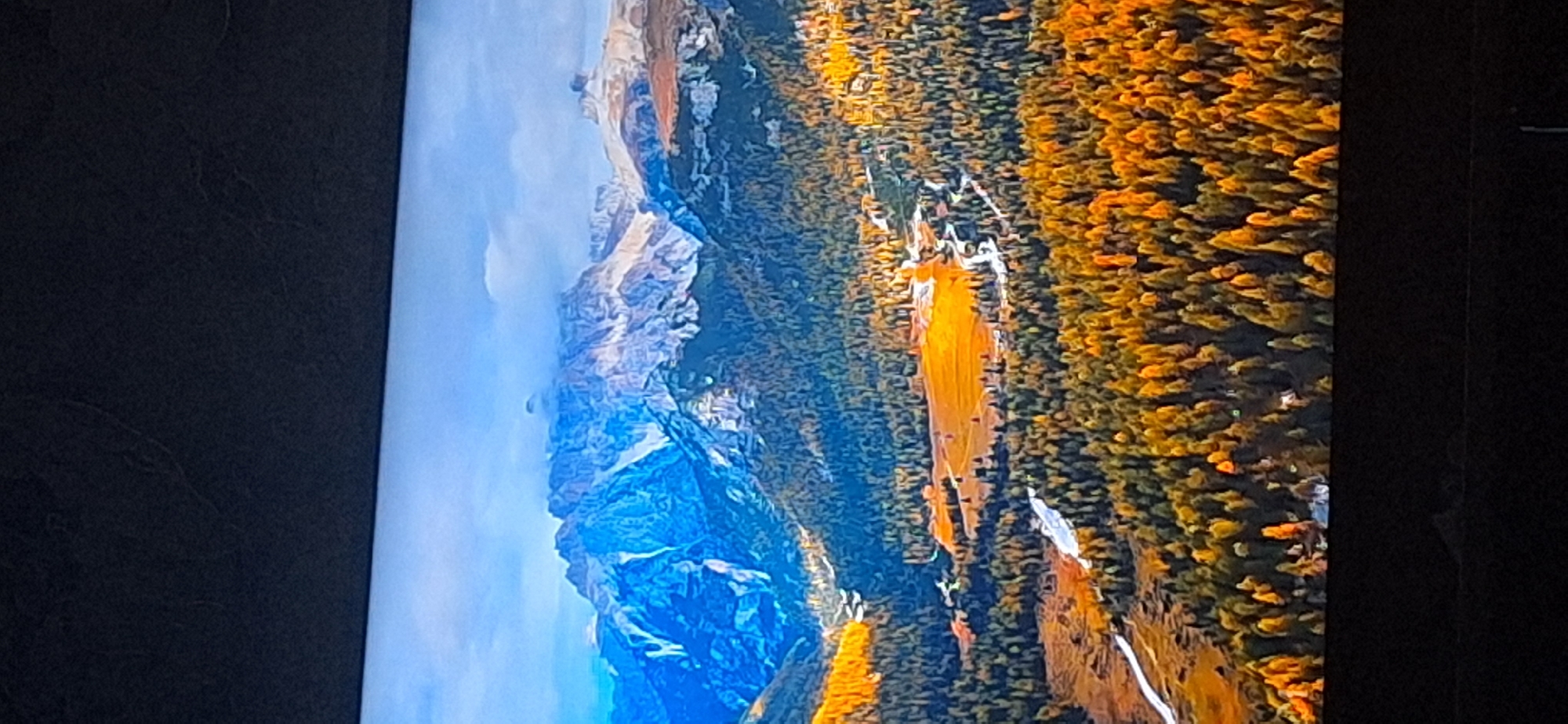 The image displays a majestic mountain landscape during autumn. The peaks are rugged and snow-capped in some areas, with dramatic cliffs and valleys. The lower slopes are covered in dense forests, primarily evergreen trees interspersed with deciduous trees displaying vibrant fall foliage in shades of yellow, orange, and brown. A winding river or stream is visible in the foreground, flowing through a valley. In the middle ground, there is a clearing or meadow with a warm, golden hue, possibly due to the sunlight hitting autumn grasses or crops. The sky is partly cloudy, with some areas of blue sky visible. The overall scene evokes a sense of natural beauty and the grandeur of the mountains in the fall season. The provided image does not contain any elements that would allow for identification of the location within the city of Bila Tserkva, Ukraine, as it depicts a natural mountain environment.