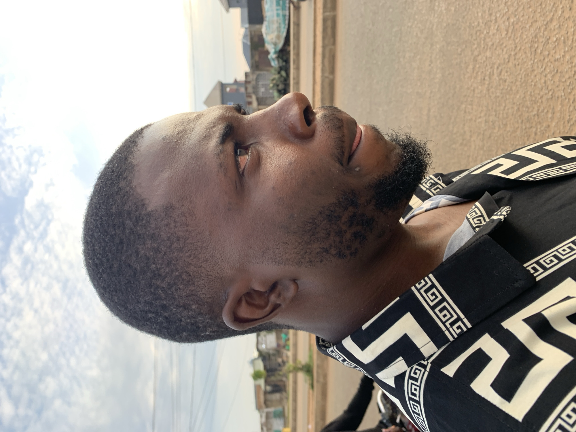 This is a medium shot, profile photograph of a Black man in Rafin Par, Nigeria. The man is positioned on the right side of the frame, his head and torso facing right. He has short, dark, curly hair, a light beard, and is wearing a black shirt with a white geometric pattern. His mouth is slightly open, and his tongue is visible. In the background, there is a paved road with a sidewalk and some buildings, indicating an urban environment. The sky is partly cloudy.