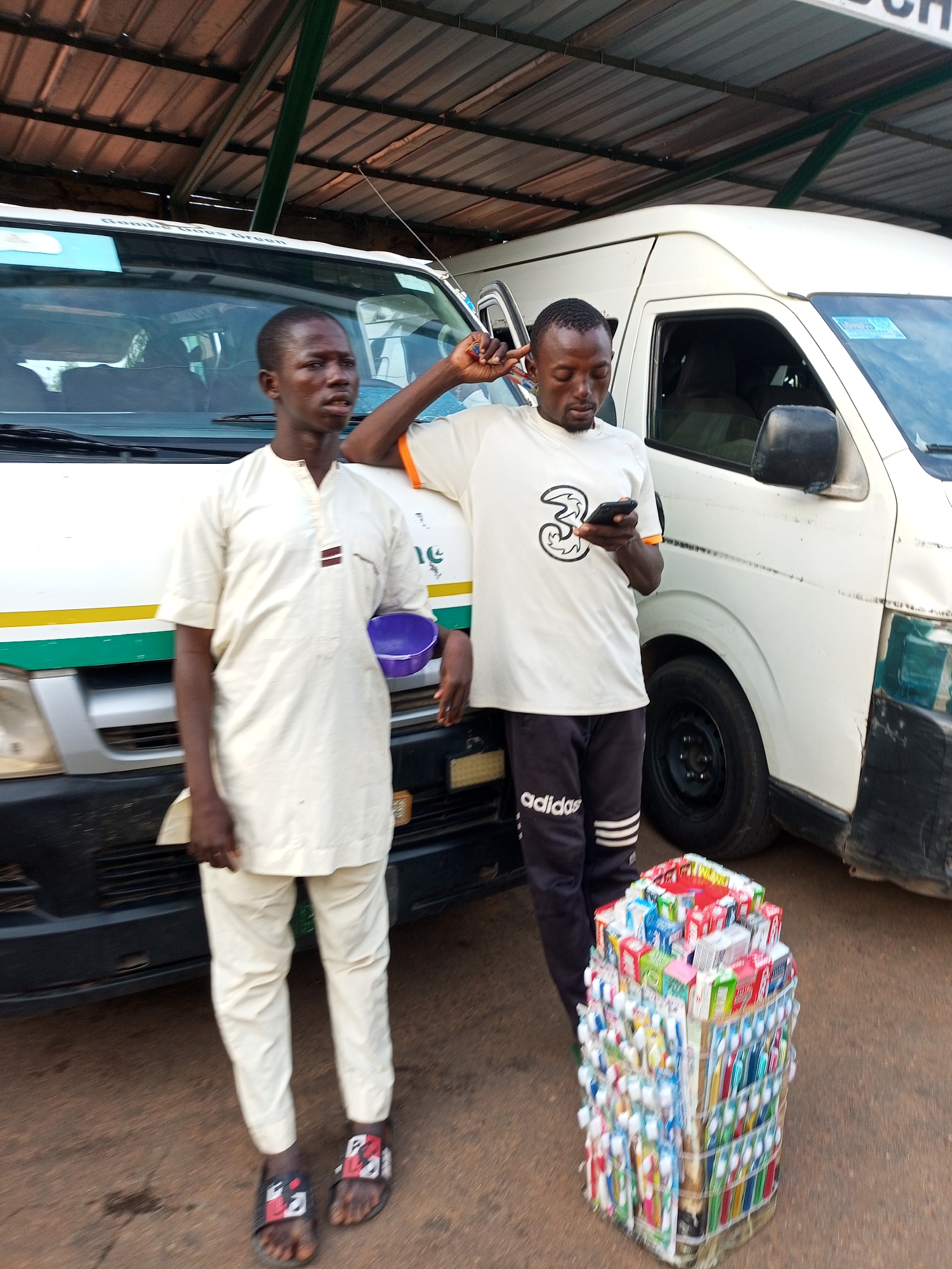 Two men are standing near a white commercial van under a covered structure. The man on the left is wearing a cream-colored tunic and trousers, and he is holding a purple bowl. The man on the right is wearing a white t-shirt with a black design and dark athletic pants with white stripes. He is looking at his phone. In front of the man on the right is a tall display of toothbrushes and toothpaste. The ground is dirt or gravel.