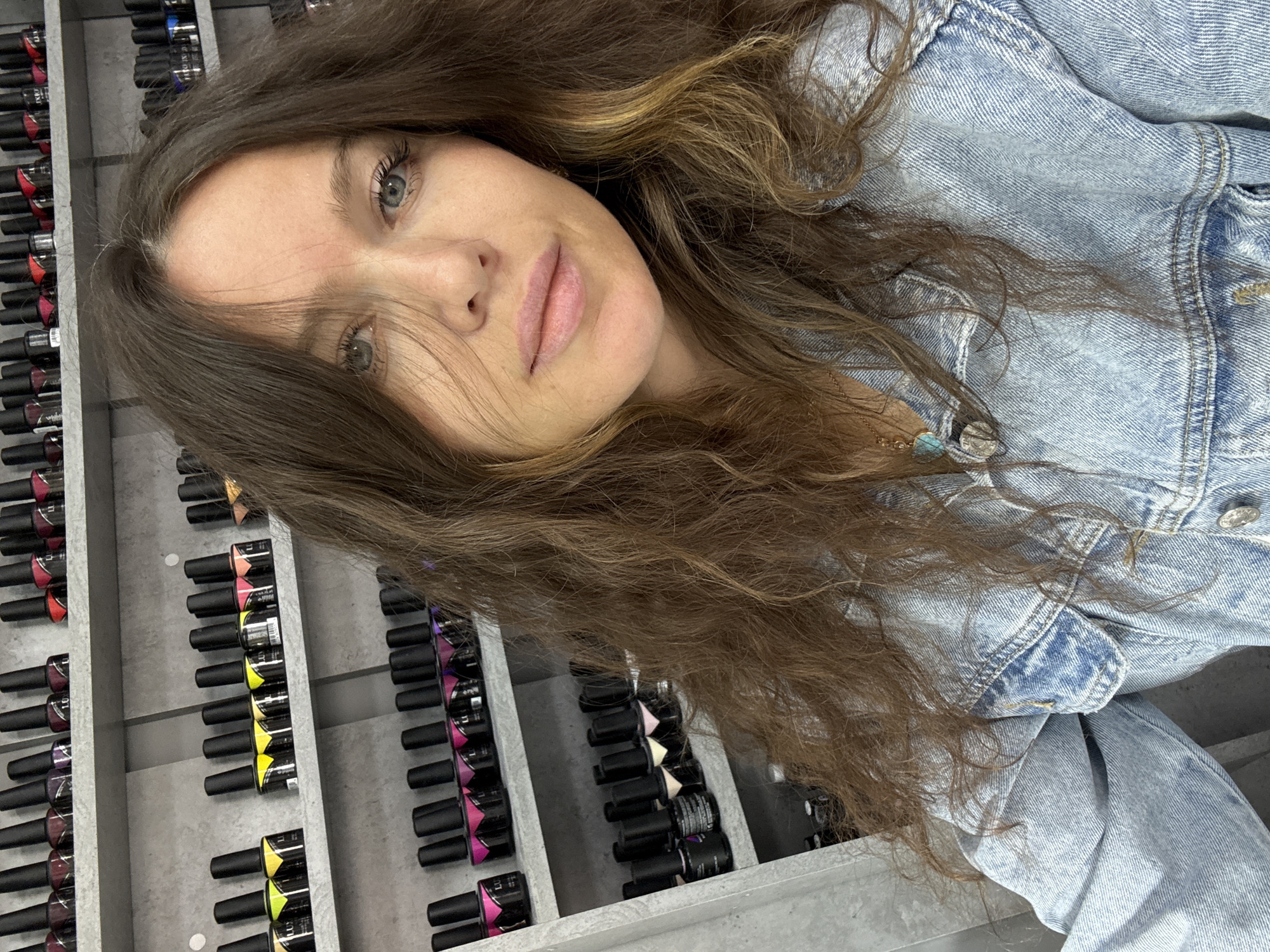 A tilted selfie portrait captures a young woman indoors, likely within a nail salon or beauty supply store located in Moscow, Russia. The woman, with long, wavy, medium-brown hair featuring lighter caramel highlights, looks directly at the camera with light blue-green eyes. Her hair cascades over her shoulders and part of her face, with a calm, slightly smiling expression on her lips. She is dressed in a light blue denim jacket, and a delicate gold chain necklace with a small turquoise and another circular charm is visible around her neck.
The background is dominated by extensive displays of nail polish bottles, arranged neatly in rows on gray shelving units. Hundreds of small bottles with black caps and vibrant colored bases — including shades of red, pink, purple, yellow, and black — are visible, indicating a wide selection. Text on some of the bottle labels, such as "LIANAIL," "LUX," and "UV/LED," along with barcodes, is discernable. The image is framed at an angle, giving a dynamic, casual feel to the scene, highlighting the woman in her commercial beauty product surroundings. The time of day is not evident, as the setting is illuminated by indoor lighting.