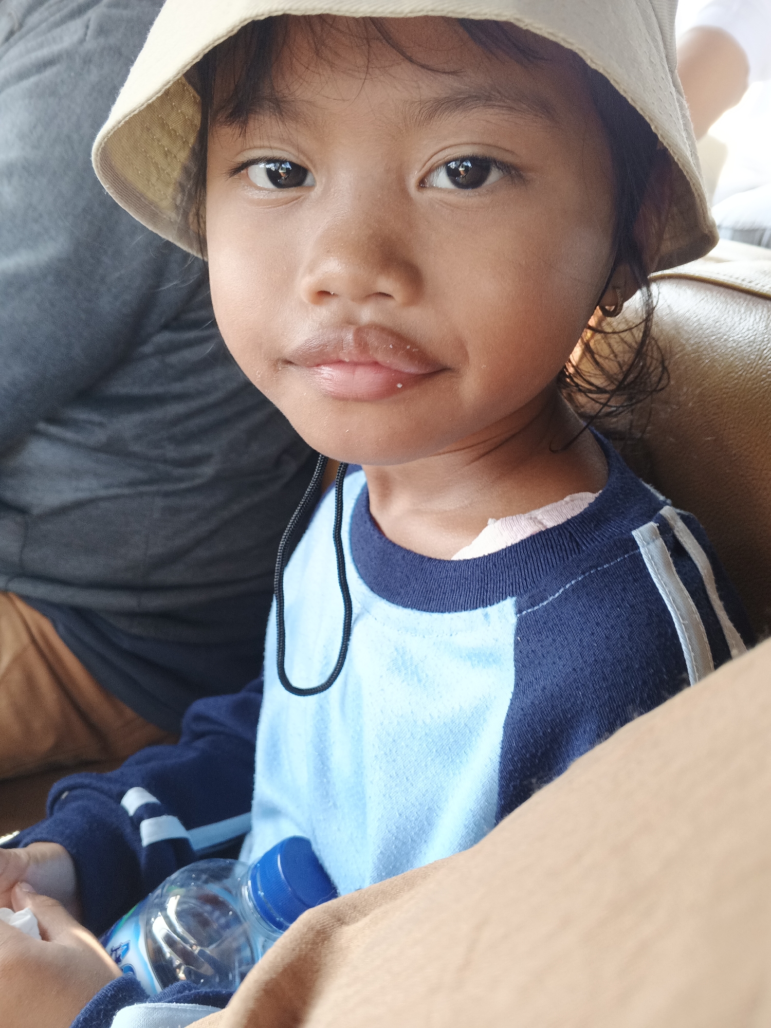A young girl wearing a light-colored bucket hat and a blue and light blue long-sleeved shirt is shown in a close-up portrait. She is sitting in what appears to be a vehicle, with brown upholstery visible behind her. Her head is tilted slightly to her right, and she is looking directly at the camera. A dark cord hangs down from her collar. In the lower left portion of the image, a partially visible plastic water bottle with a blue cap is present. The background is blurred, suggesting a shallow depth of field. The lighting indicates it is daytime. The image appears to have been taken in Sukabumi, Indonesia.