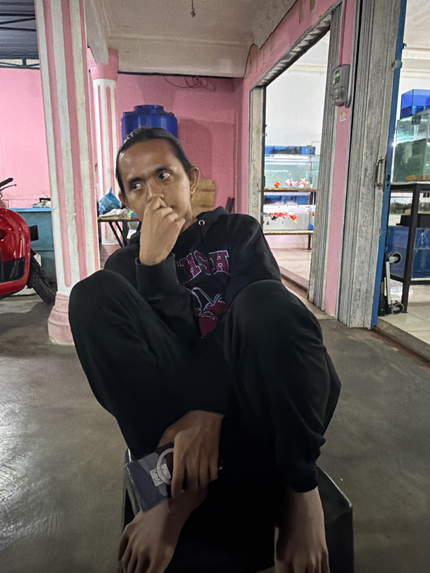The image shows a person sitting on a chair in what appears to be an outdoor area, possibly a shop or a market, in Tanjung Pinang, Indonesia. The background features pink walls with white striped pillars, a red motorcycle on the left, and several fish tanks with goldfish in the background to the right. The person is wearing a black hoodie and black pants, with their legs crossed and resting on the chair. They are holding a mobile phone in their hand and have their other hand on their chin, looking to the side with a contemplative expression. The floor is concrete. The lighting suggests it is either early morning or late evening.