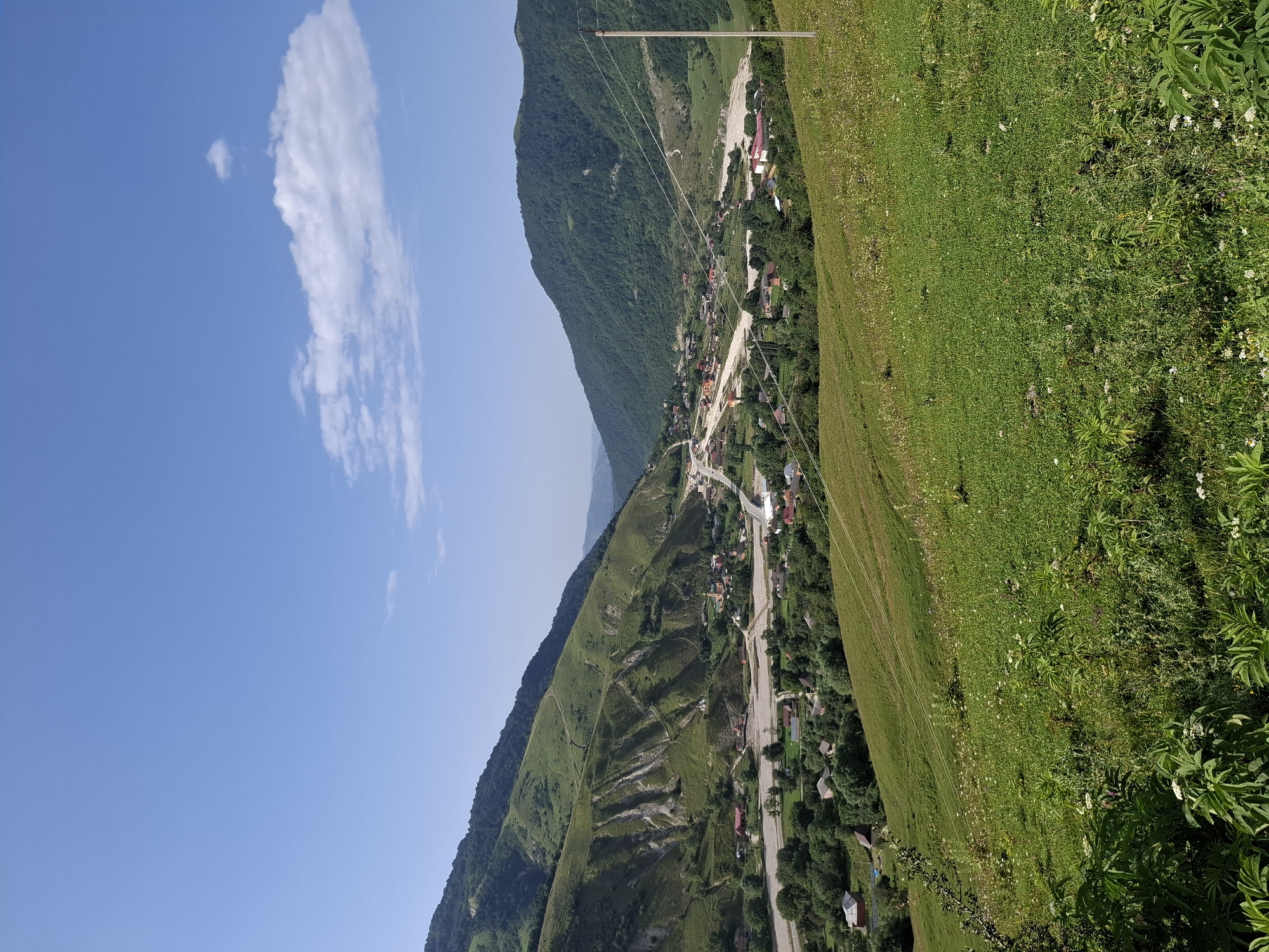The image captures a panoramic view of a village nestled within a valley in Kharachoy, Russia. Lush green hills flank both sides of the valley, with the slopes on the left exhibiting signs of erosion and bare patches, while the right side is densely forested. A riverbed, appearing dry or with minimal water flow, cuts through the valley floor, with a road winding alongside it. Scattered throughout the valley are numerous houses with red and brown roofs, indicating a residential area. In the foreground, a grassy hillside slopes downward, dotted with wildflowers. The sky is a clear blue with a few scattered white clouds, and a power line stretches across the scene from a wooden pole on the right. The overall impression is a tranquil rural landscape under a bright, sunny sky.