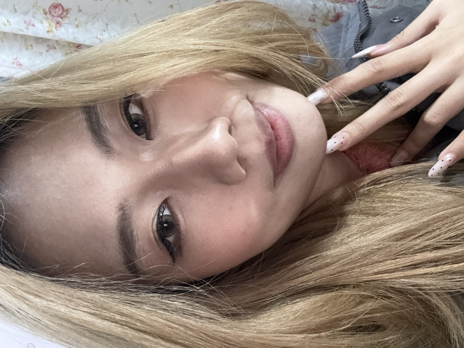 An indoor, close-up portrait shot, presumably a selfie, depicts a female individual in Shapingba District, China. The subject, appearing to be of East Asian ethnicity, has fair skin and long, straight, light blonde hair that frames her face and extends over her shoulders. Her eyes are open, revealing dark brown irises, possibly enhanced with contact lenses, and precise black eyeliner. Her eyebrows are well-defined. Her lips are slightly parted in a subtle smile, tinted with a light pink color, and her cheeks show light blush. The individual's left hand is positioned with the fingertips resting against her chin. This hand displays long, almond-shaped fingernails, painted white with small black polka dots. She wears a reddish undergarment visible beneath a grey outer layer of clothing. The background is shallowly focused, showing a floral patterned curtain with pink and green motifs on the right side of the frame. The scene is evenly lit.