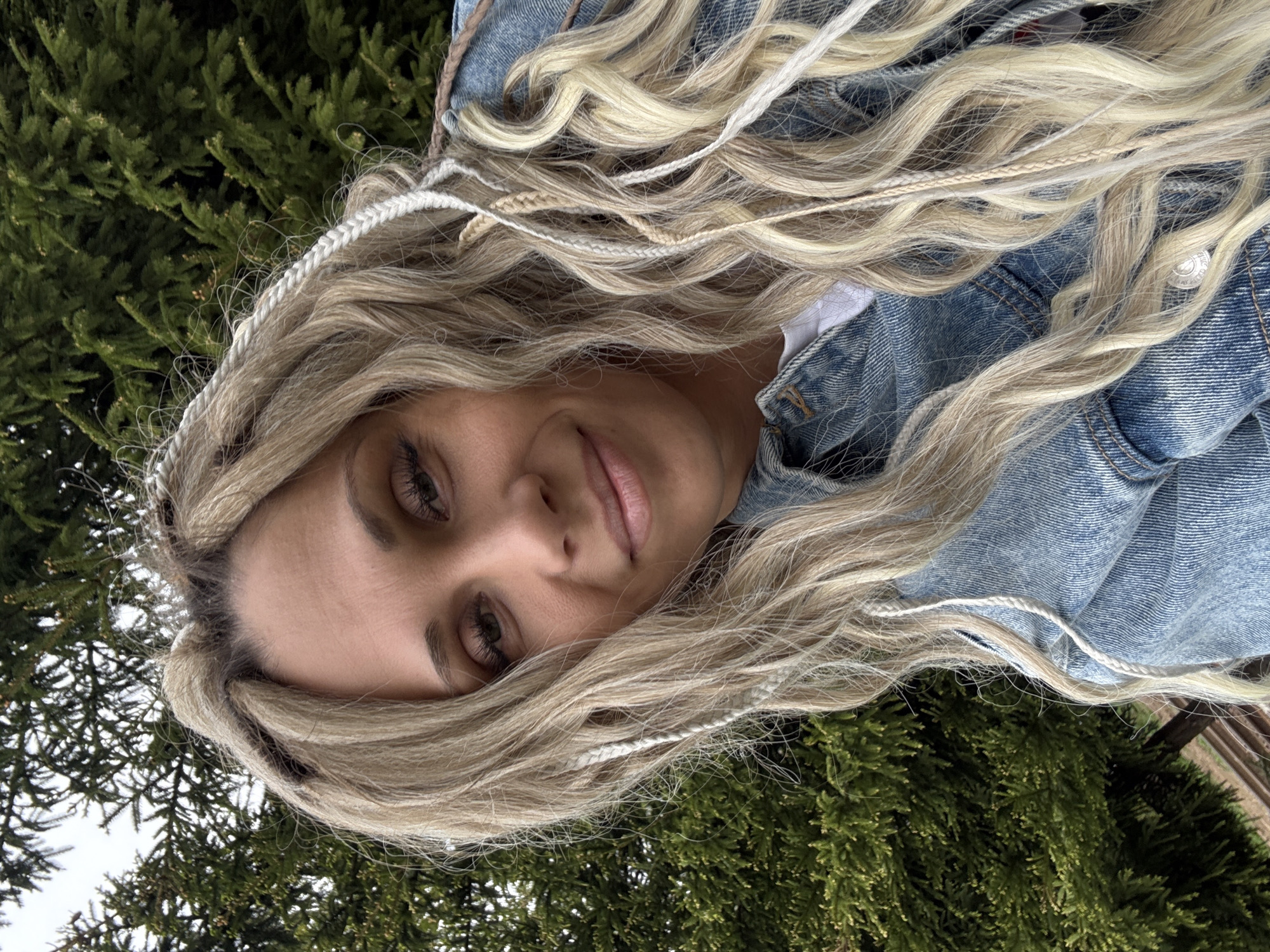 A woman with long, blonde wavy hair interwoven with white braids is positioned in front of dense green foliage, possibly a coniferous bush or small trees. She is wearing a denim jacket and appears to be looking directly at the camera. The setting suggests an outdoor location, potentially a park or garden. There is no specific information to confirm the location as Rusanovo, Russia from the image.
