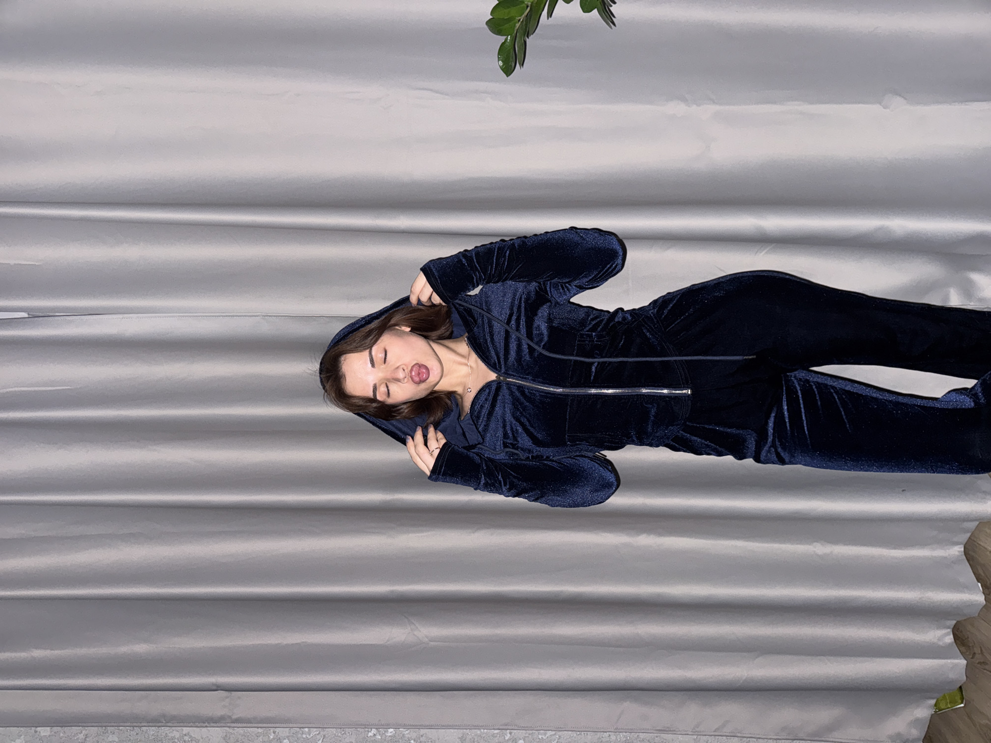 A woman wearing a navy blue velvet tracksuit is lying on a gray, horizontally-pleated fabric surface. The woman has light skin and brown hair, and her mouth is open with her tongue slightly extended. She is wearing a hooded sweatshirt zipped halfway down, and a necklace is visible around her neck. A green leafy branch is visible at the top of the image. The background texture and lighting suggest an indoor setting, potentially a staged photoshoot in a studio. The exact location in Лобаново, Russia, is not discernible from the image.