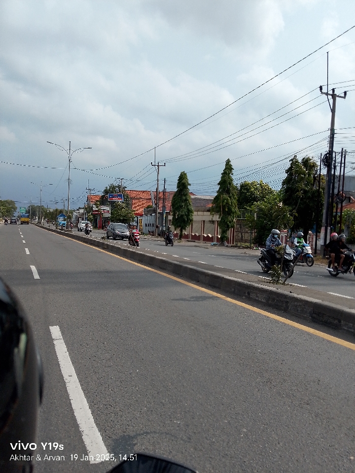 The image captures a perspective from a vehicle, likely a motorcycle, on a busy road in Arjawinangun, Indonesia, on an overcast afternoon. A portion of a black helmet and a rearview mirror are visible in the lower left foreground, suggesting the viewpoint.
The scene features a wide asphalt road divided by a concrete median with a yellow curb line. White dashed lines mark the lanes of traffic. Numerous motorcycles and scooters are actively using the road, with several riders visible, some wearing helmets. A dark grey car is seen on the left side of the median, and a yellow bus or truck is discernable in the far distance.
Along both sides of the road, particularly to the right, are buildings with distinctive orange tiled roofs, suggesting a mix of residential and commercial structures. Tall utility poles with a dense network of overhead electrical or communication wires line the street. Several tall, slender evergreen trees and other leafy trees are planted alongside the buildings and the road, providing greenery.
The sky is heavily overcast with significant cloud cover, indicating a cloudy day. The time of the photo is precisely indicated by the visible text "19 Jan 2025, 14.51" (2:51 PM). Other text visible in the bottom left corner includes "vivo Y19s," identifying the camera model, and "Akhtar & Arvan," likely names associated with the photograph. The overall activity depicts routine daily traffic in an urban or semi-urban Indonesian setting.