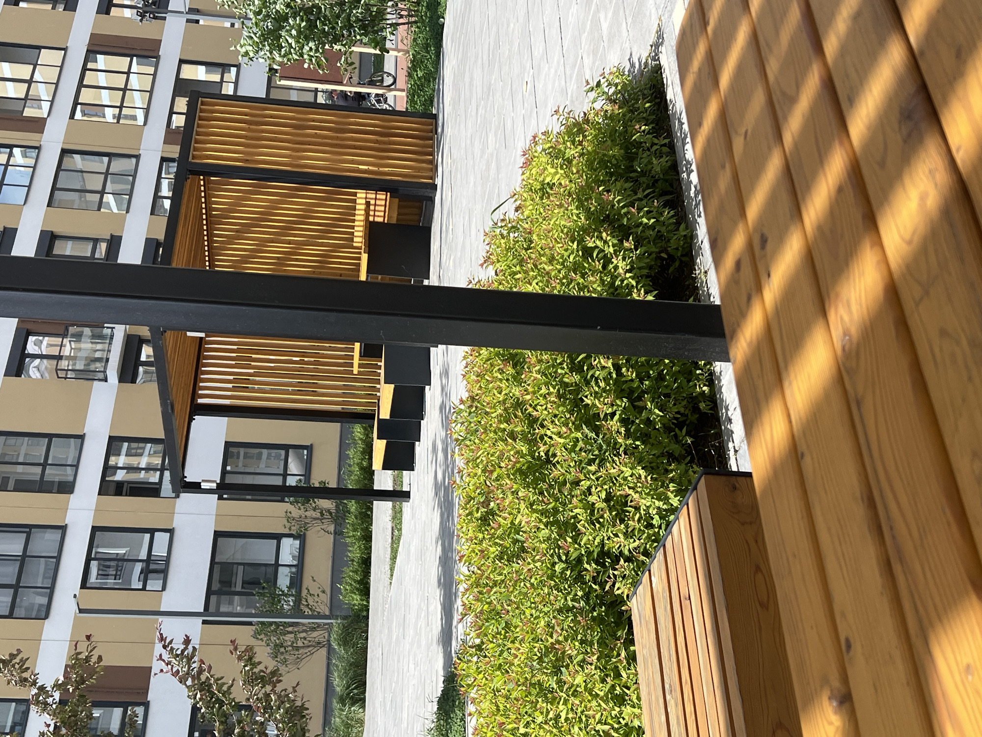 A photograph taken in Yekaterinburg, Russia, shows an outdoor urban space. The scene features modern architecture with a multistory building on the left, characterized by multiple rows of rectangular windows. The building's facade is a combination of light tan and white. In the foreground, there is a wooden bench running along the right side of the frame. Next to the bench, there is a strip of green bushes. A paved walkway runs between the bushes and a series of open wooden structures with black metal frames, positioned in the midground to the left. The structures appear to be shaded sitting areas. The photograph is shot from a close perspective, with some elements in the foreground slightly blurred, drawing attention to the details in the midground.
