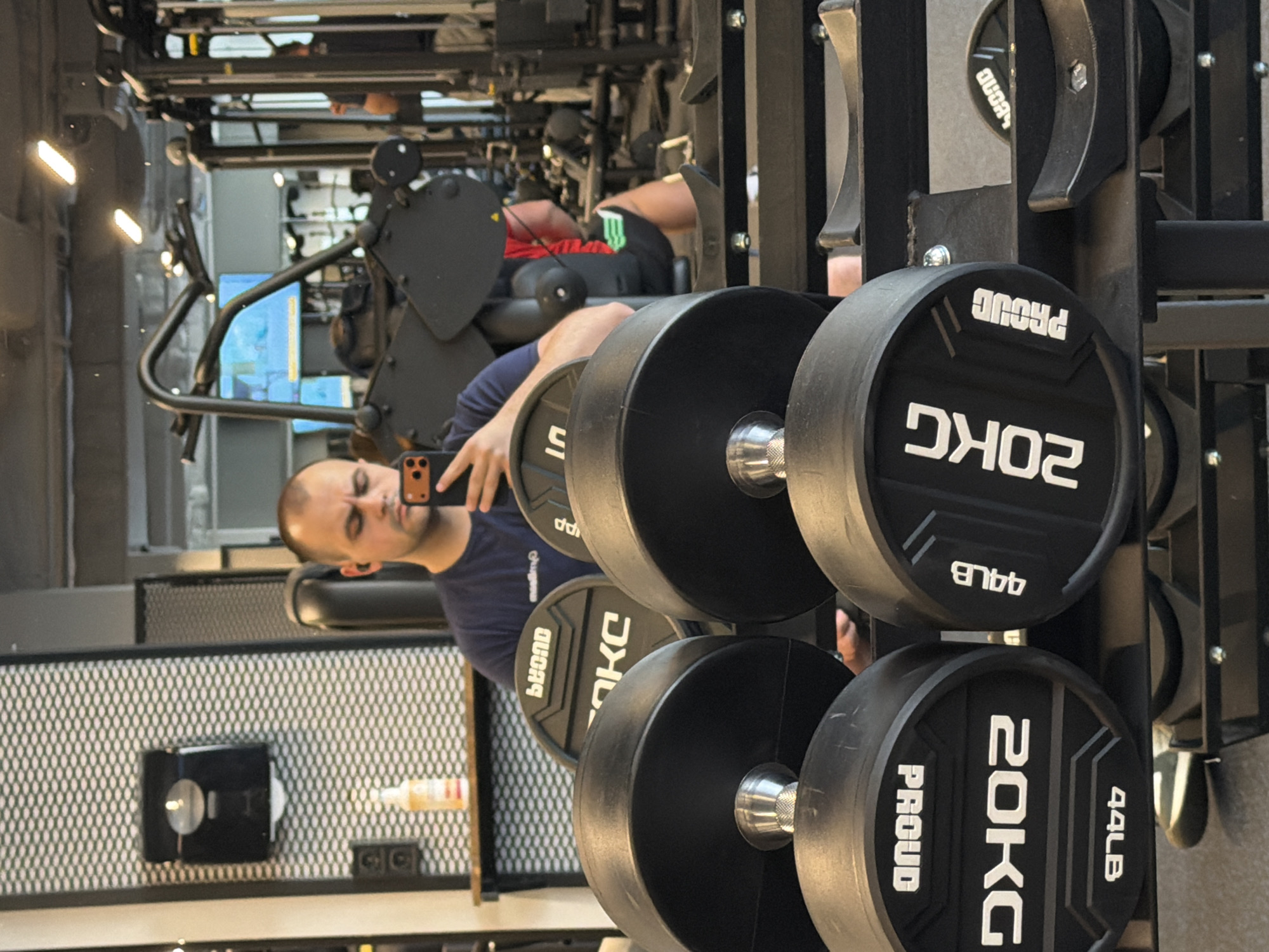 A mirror in a gym in Plovdiv, Bulgaria, reflects a man taking a selfie with his phone. To his right, a weight rack holds 20 KG "PROUD" weights. In the background, another person is using a weight machine. A water cooler, outlets, and a perforated wall are visible behind the man in the reflection.