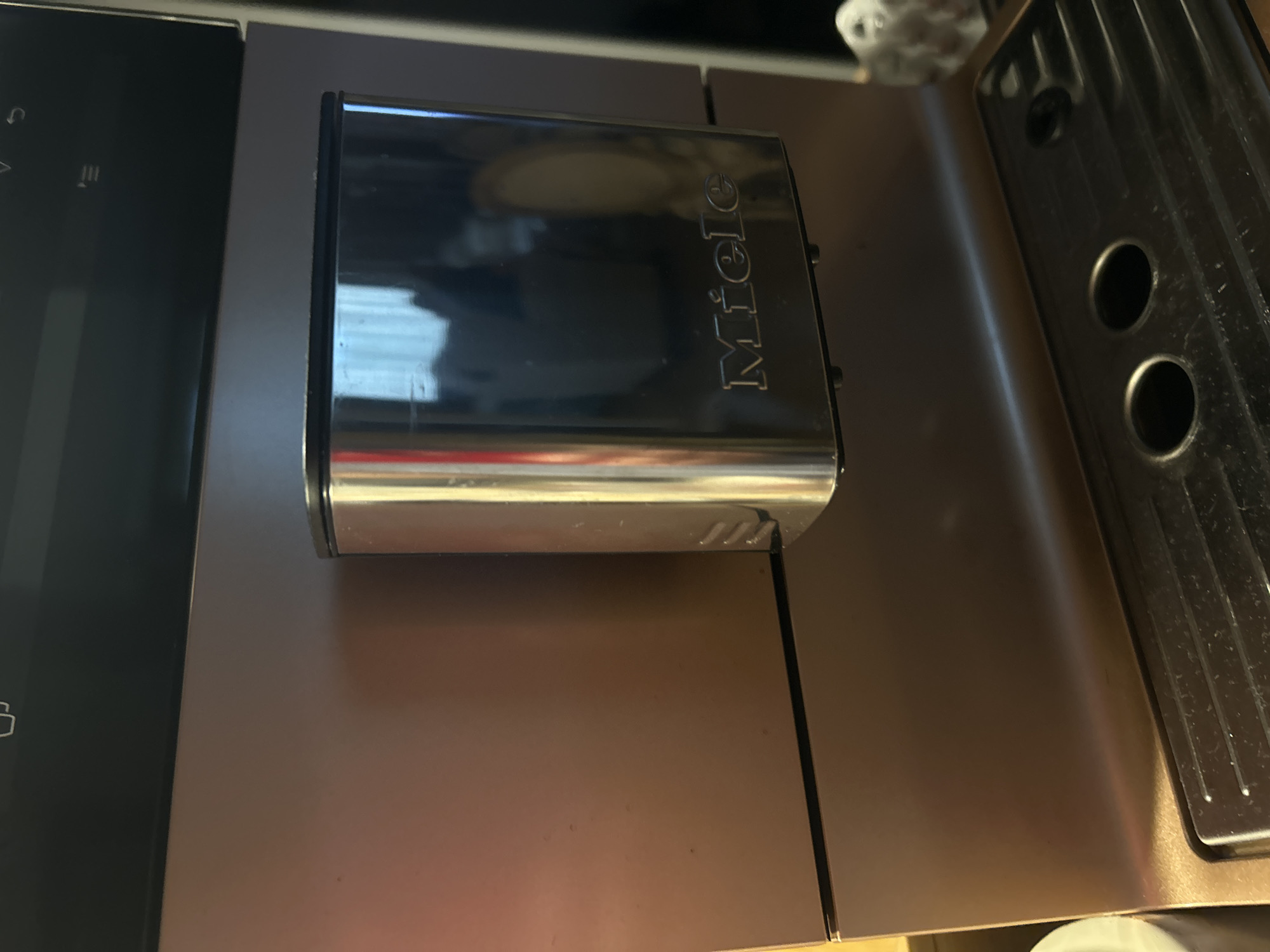 The image shows a close-up of a Miele coffee machine. The brand name "Miele" is visible on a shiny, chrome-colored rectangular component. To the left is part of the control panel. To the right is a section with circular holes. The machine appears to be sitting on a brown or bronze-colored surface. The location is not apparent from the image.