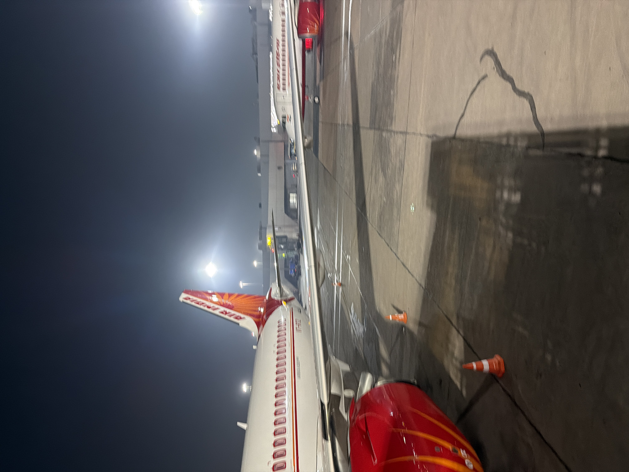 An Air India airplane is parked on the tarmac at Indira Gandhi International Airport in New Delhi, India. The aircraft's tail, emblazoned with the Air India logo, is visible in the upper right portion of the frame. The main body of the plane, featuring rows of windows and the registration number VT-ATD, runs horizontally across the image. To the left, a red engine cowling is prominent. The scene takes place at night, indicated by the artificial lighting illuminating the airport environment. Several orange traffic cones are placed on the tarmac, likely for safety or guidance. The ground is concrete, with visible seams and some dark markings or shadows. In the background, airport infrastructure, including what appears to be a jet bridge and other aircraft, is dimly lit. The sky is dark, with some bright lights from the airport.