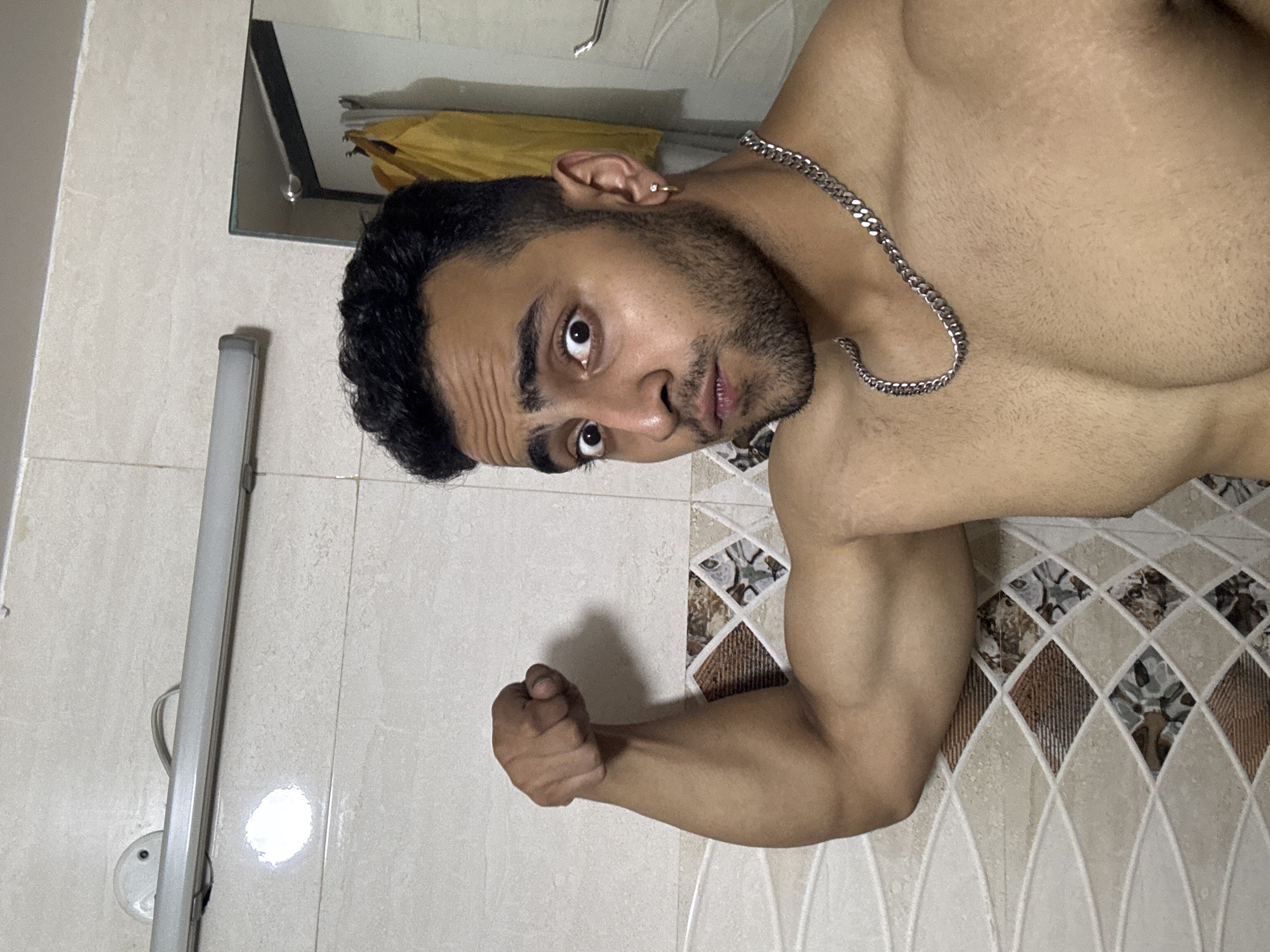 A shirtless South Asian man in his mid-20s is posing in a bathroom. He has short, dark hair and a trimmed beard. He is wearing a silver chain necklace and a small hoop earring in his left ear. His right arm is flexed, showcasing his bicep. The bathroom has off-white and brown patterned tiles. There is a light fixture and a mirror on the wall behind him. The city of Kolkata, India is not discernible from the image.
