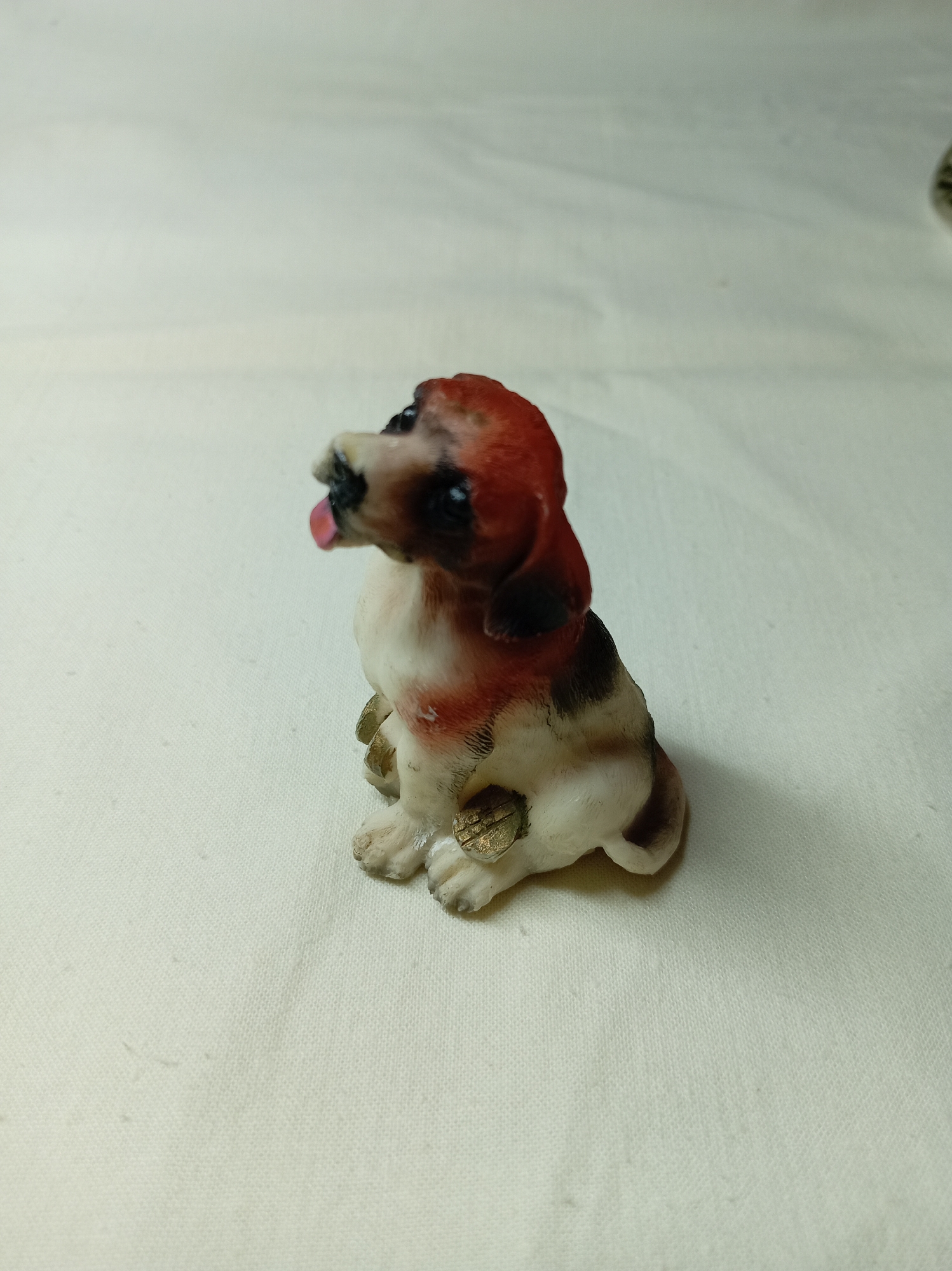 A close-up shot features a small, decorative figurine of a dog. The dog is seated, with a predominantly white body marked with brown and red patches. It has dark eyes and a visible pink tongue. Small, golden-colored, scale-textured objects are near its paws. The figurine is positioned on a white, slightly creased surface, possibly a cloth or sheet. The image lacks any specific visual markers that confirm a location in Bugulma, Russia; therefore, the location is not discernable from the image alone.