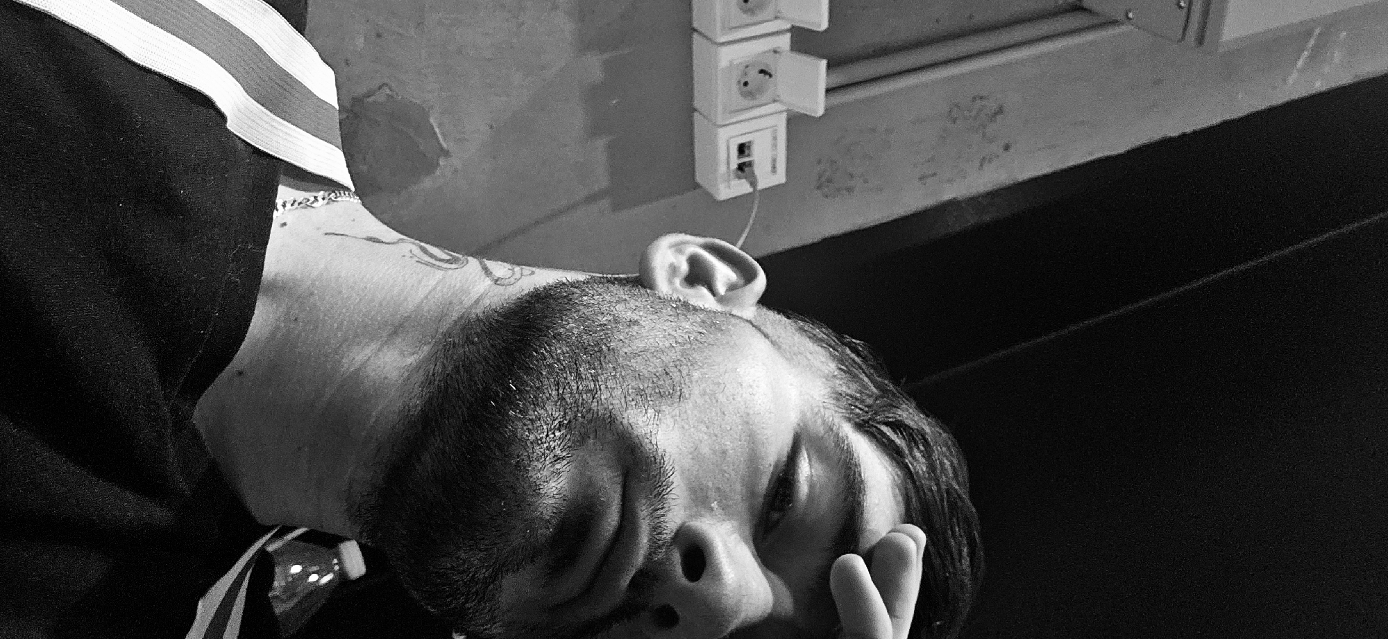 A black and white image shows a man with a shaved head and a small tattoo on his neck. He is lying down with his eyes closed and his hand near his face. The man is wearing a dark shirt with light-colored stripes on the shoulder. There are electrical outlets on a wall nearby. The scene is indoors. There is no information that indicates the city of Plovdiv, Bulgaria.