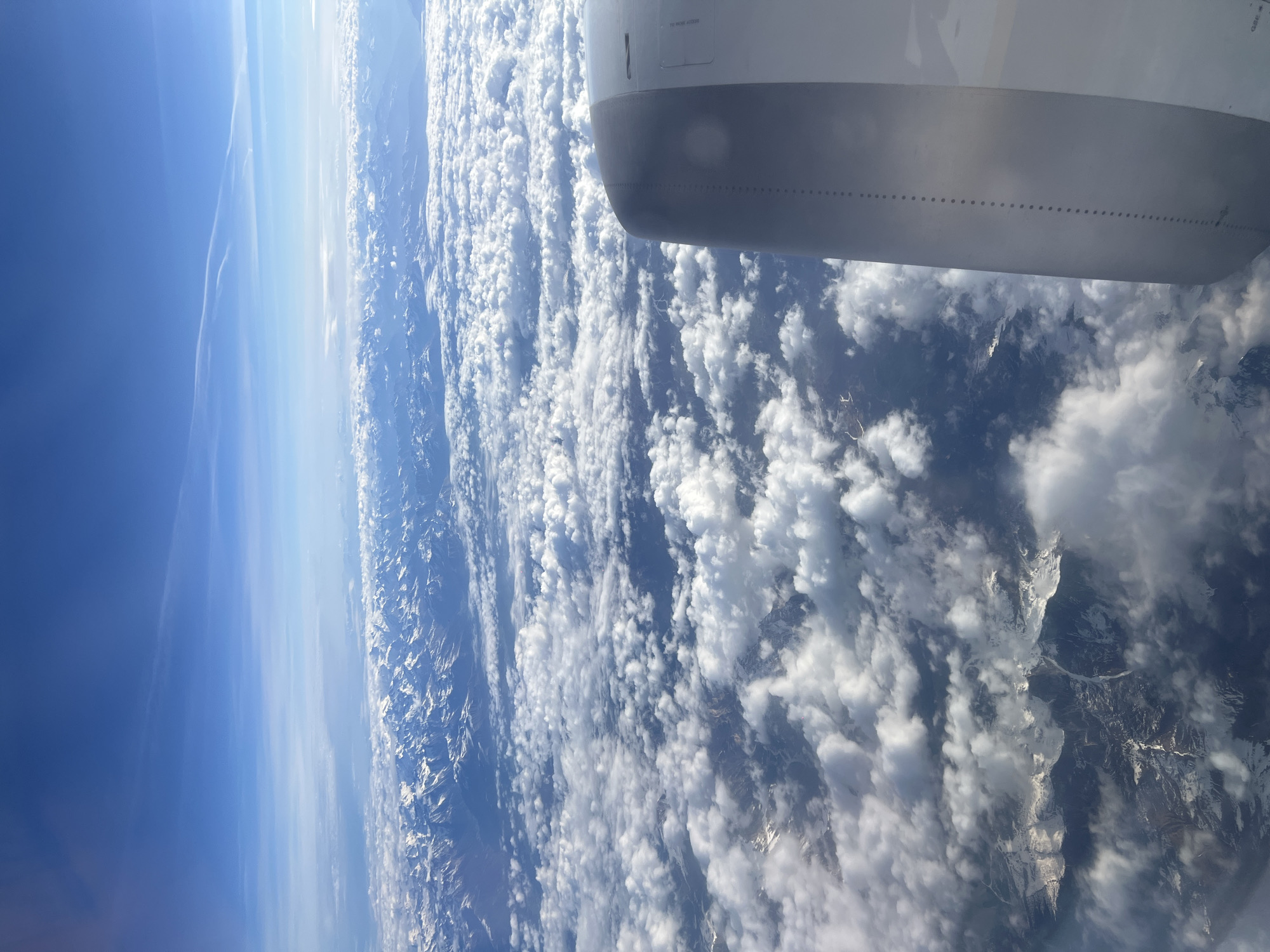 The image depicts a view from an airplane window. A portion of the aircraft wing is visible in the top right corner. Below the wing, a landscape of mountains and clouds stretches towards the horizon. The sky transitions from a deep blue to a lighter blue closer to the horizon. The mountain peaks are partially covered in snow. The land below is partially obscured by cloud cover. The location is not determinable from the image.