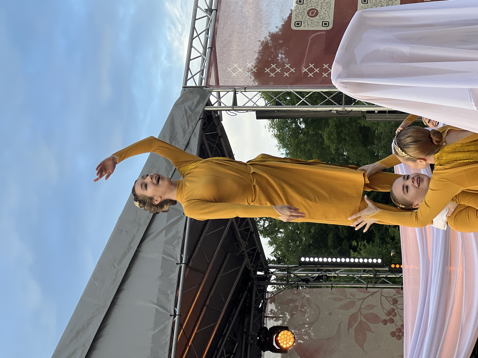 A group of young women in yellow costumes are performing on an outdoor stage. The woman at the top, held up by two others, is wearing a long-sleeved yellow dress and a gold headband. Her left arm is raised in the air, and she is looking upwards with her mouth open. The two women supporting her are also in yellow costumes, one with braces on her teeth. A third person is covered by a white cloth on the right side of the frame. The stage has lighting equipment and a backdrop with a red pattern. Trees and a cloudy sky are visible behind the stage. The performance appears to be taking place in a park or outdoor venue.