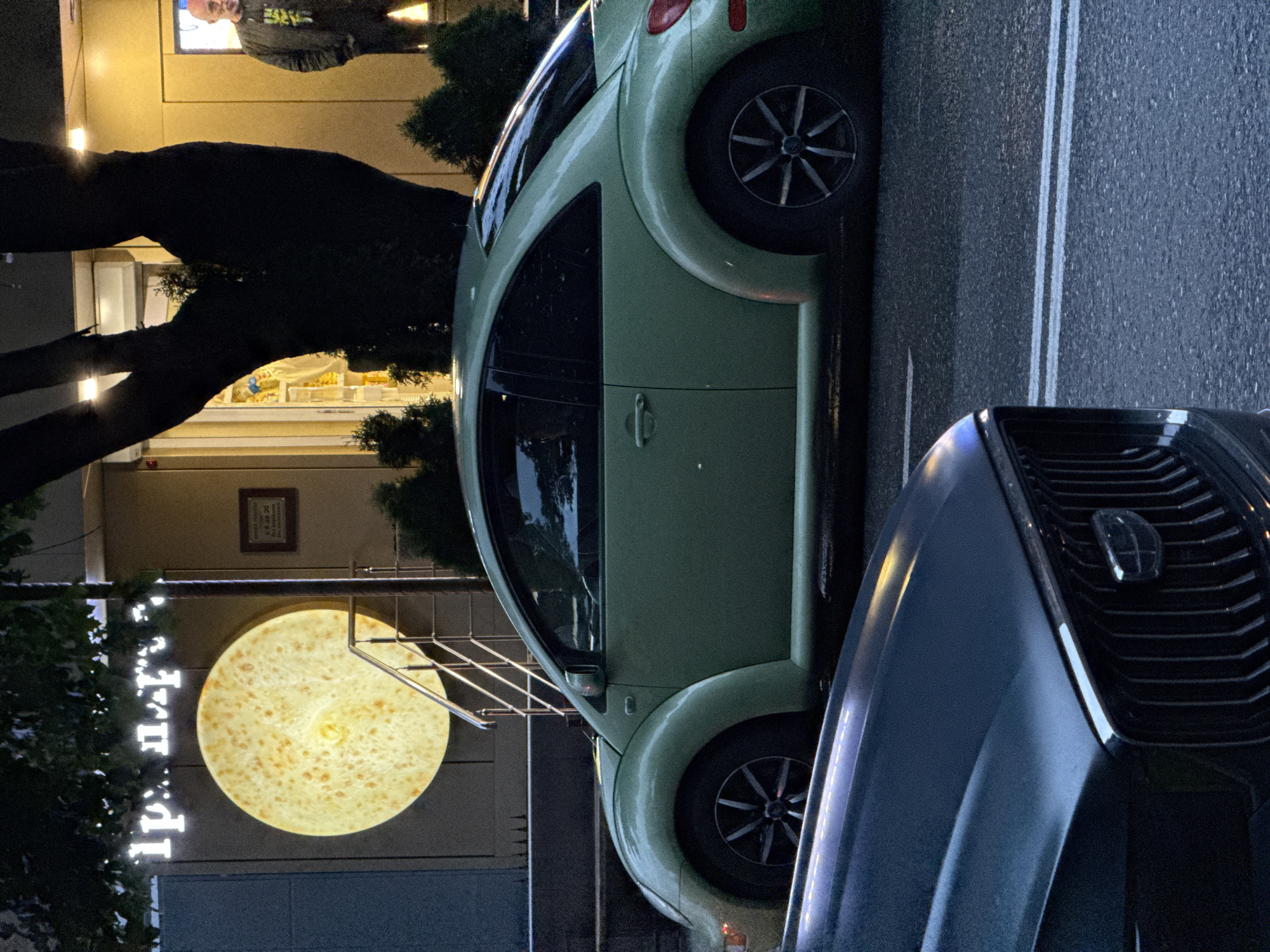 In this nighttime scene in Assinovskaya, Russia, a light green Volkswagen Beetle is parked next to another dark-colored vehicle. The Beetle's rounded silhouette is characteristic of its design. The street is paved and marked with white lane lines. To the left of the Beetle, a building with illuminated signage is visible. The sign features a large, circular, illuminated graphic resembling a flatbread or pancake, with smaller, white illuminated letters spelling out "Frudi" vertically. Above the sign, a framed plaque is mounted on the building's facade. A dark tree with sparse foliage is also present in the foreground, its branches silhouetted against the building's lights. In the background, through a window or opening in the building, a person wearing a dark jacket with yellow lettering is visible. The overall atmosphere is that of a quiet evening in a town.